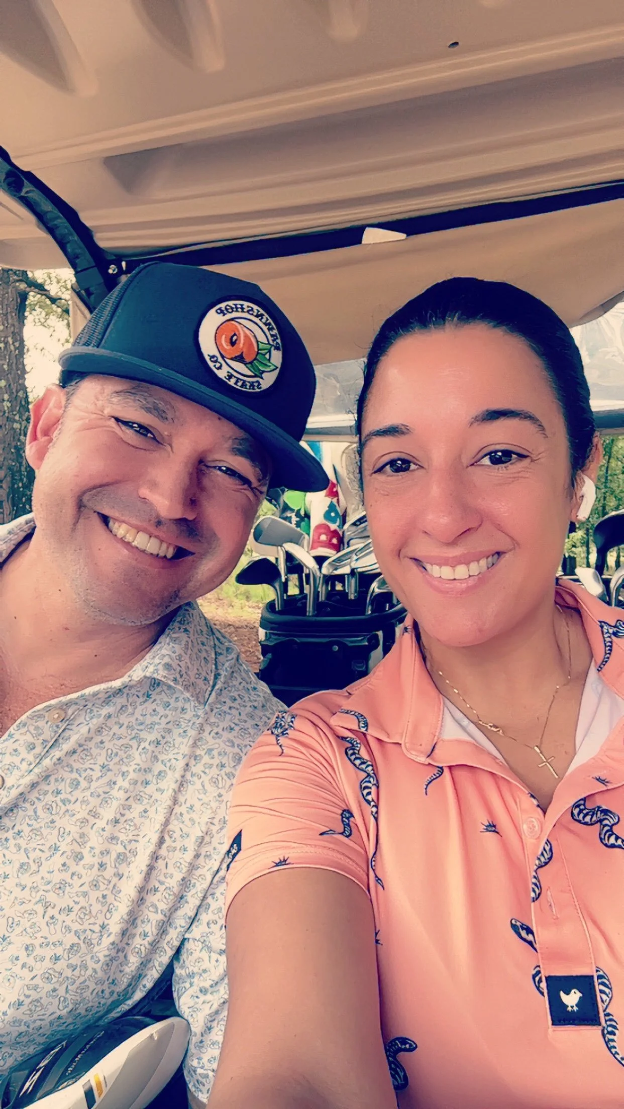 A smiling man wearing a baseball cap with a peach logo and a woman in a pink shirt with snake patterns, taking a selfie at a golf course with golf clubs in the background.