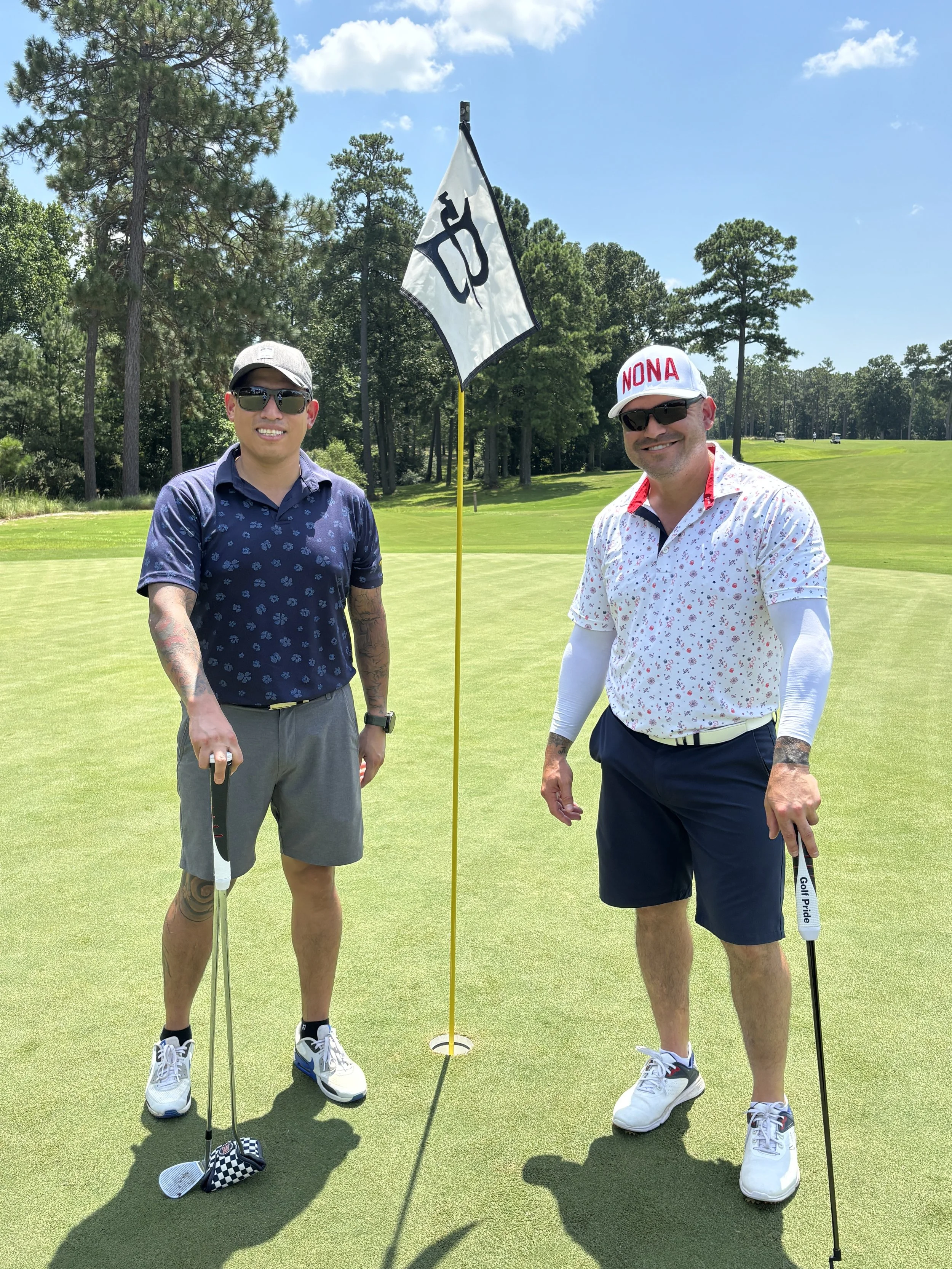 Two men standing on a golf course near a flag, holding golf clubs, wearing sunglasses, smiling, with trees and a clear blue sky in the background.