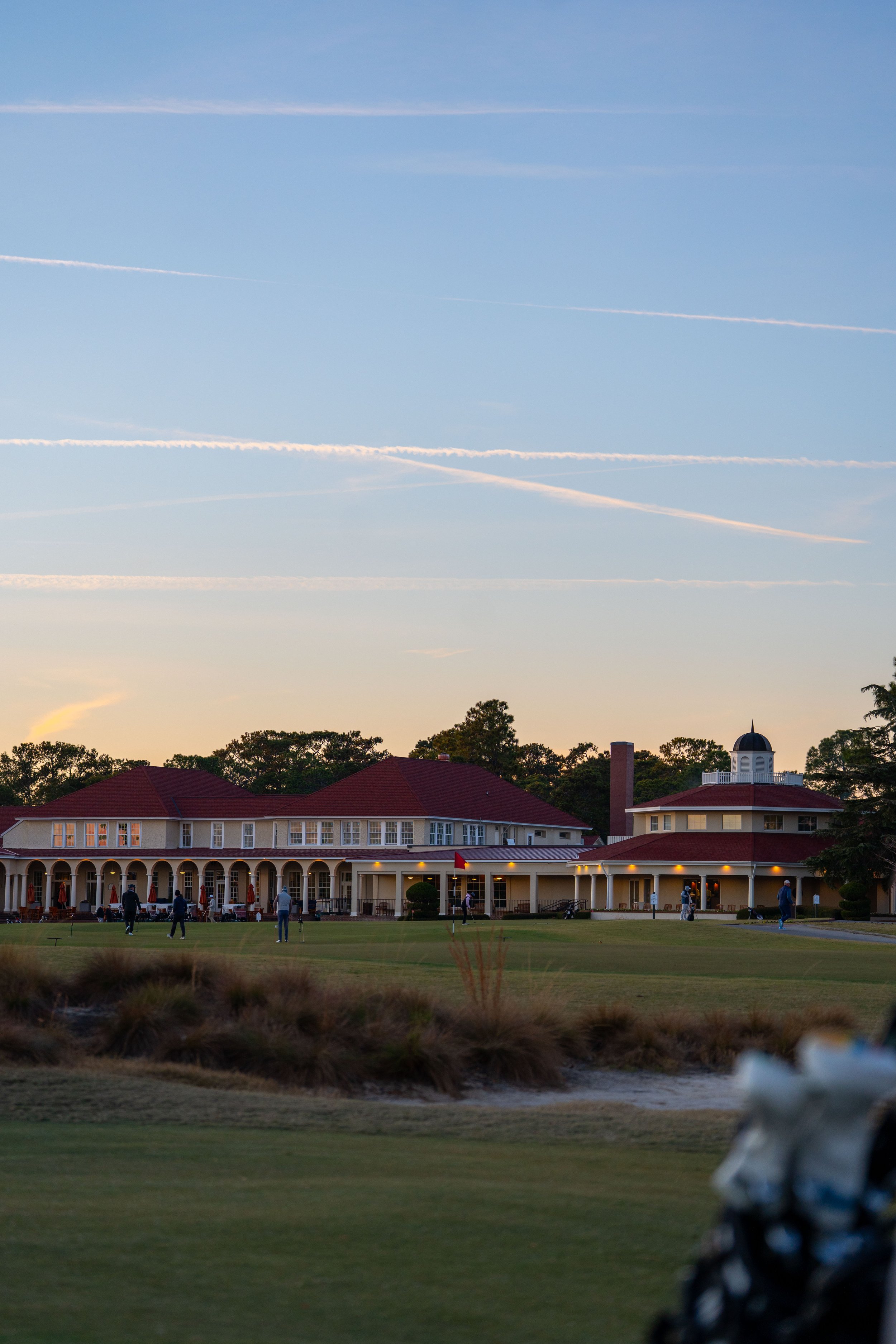 A group of people playing golf on a golf course at sunset, with a large building featuring a red roof and a clock tower in the background.