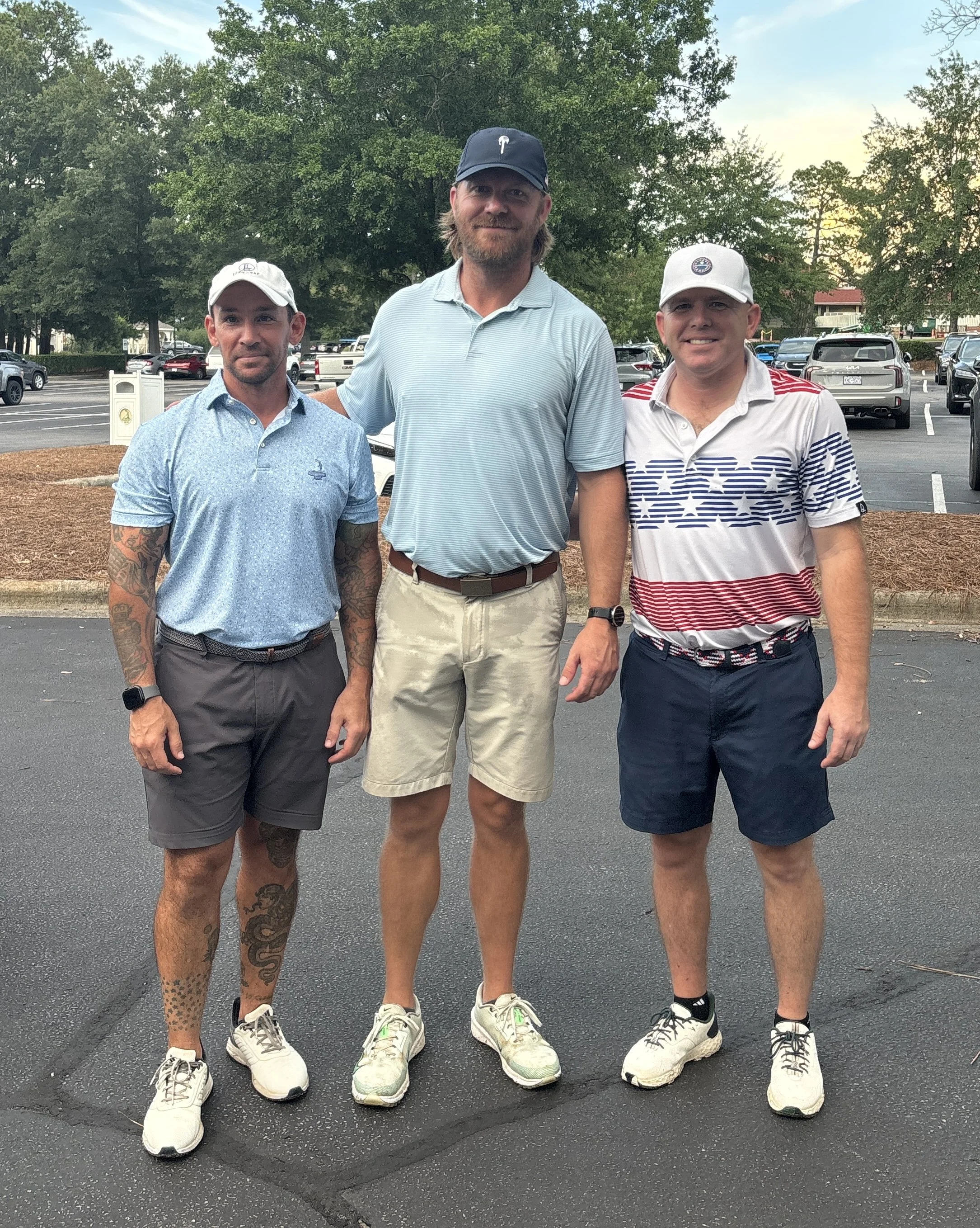 Three men standing outside on pavement in front of a parking lot with trees and cars, wearing casual golf attire.