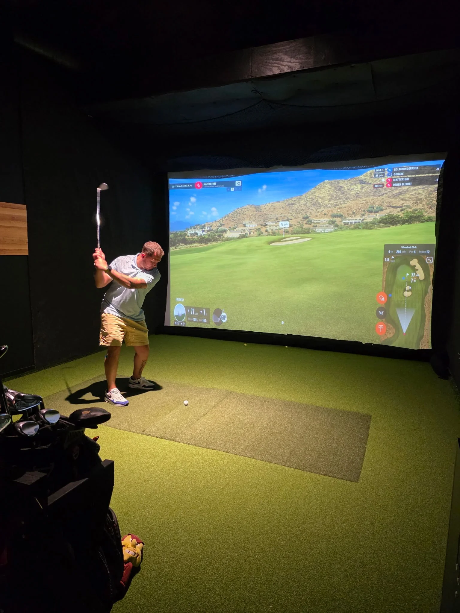 Man in a golf simulator room swinging a golf club, with a virtual golf course projected on the screen in front of him.