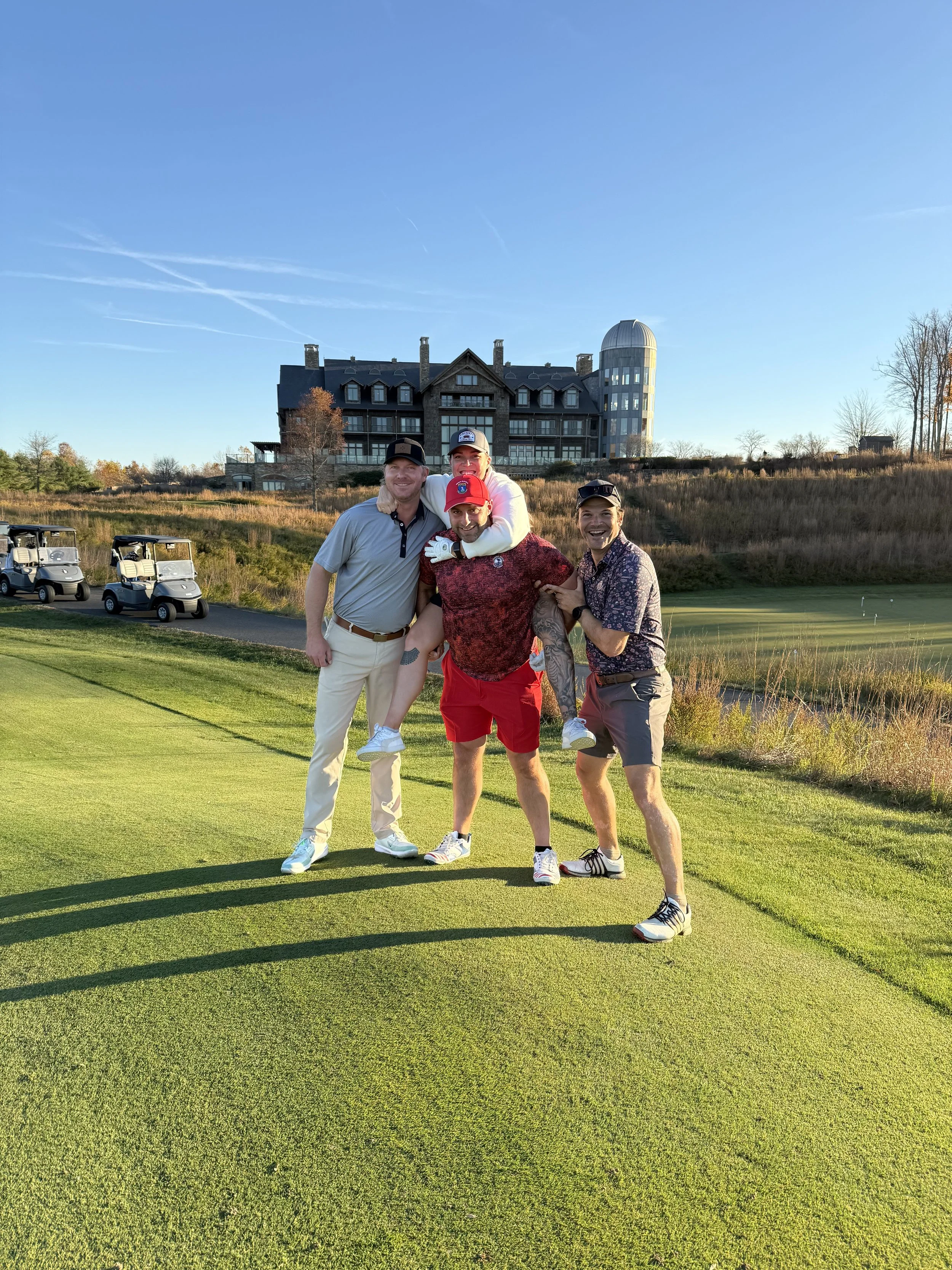 Four men standing on a golf course, smiling and posing for the photo, with a large building in the background, under a clear blue sky.