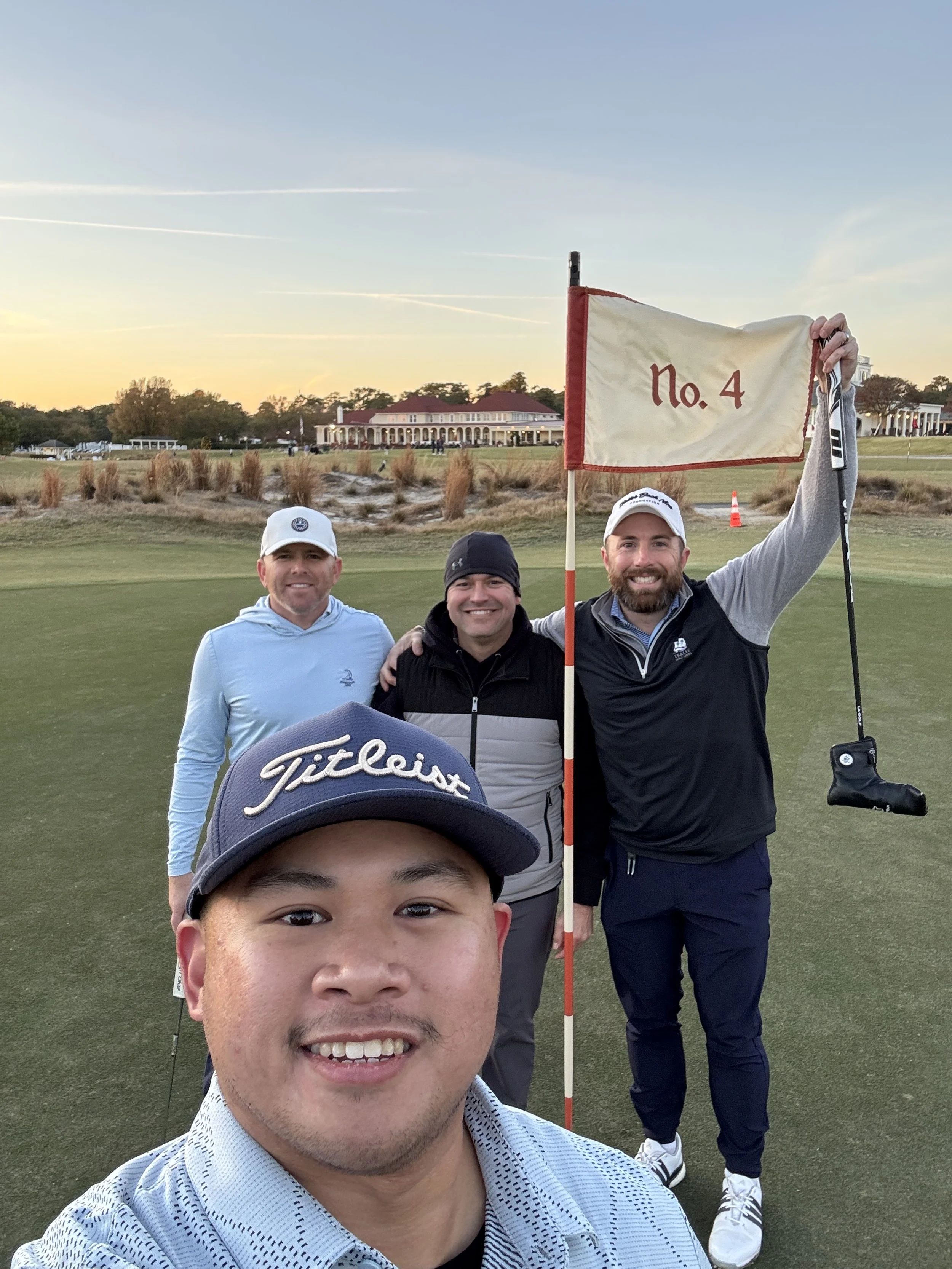 Group of four men celebrating on a golf course at sunset, one holding a flag with No. 4, smiling at the camera.