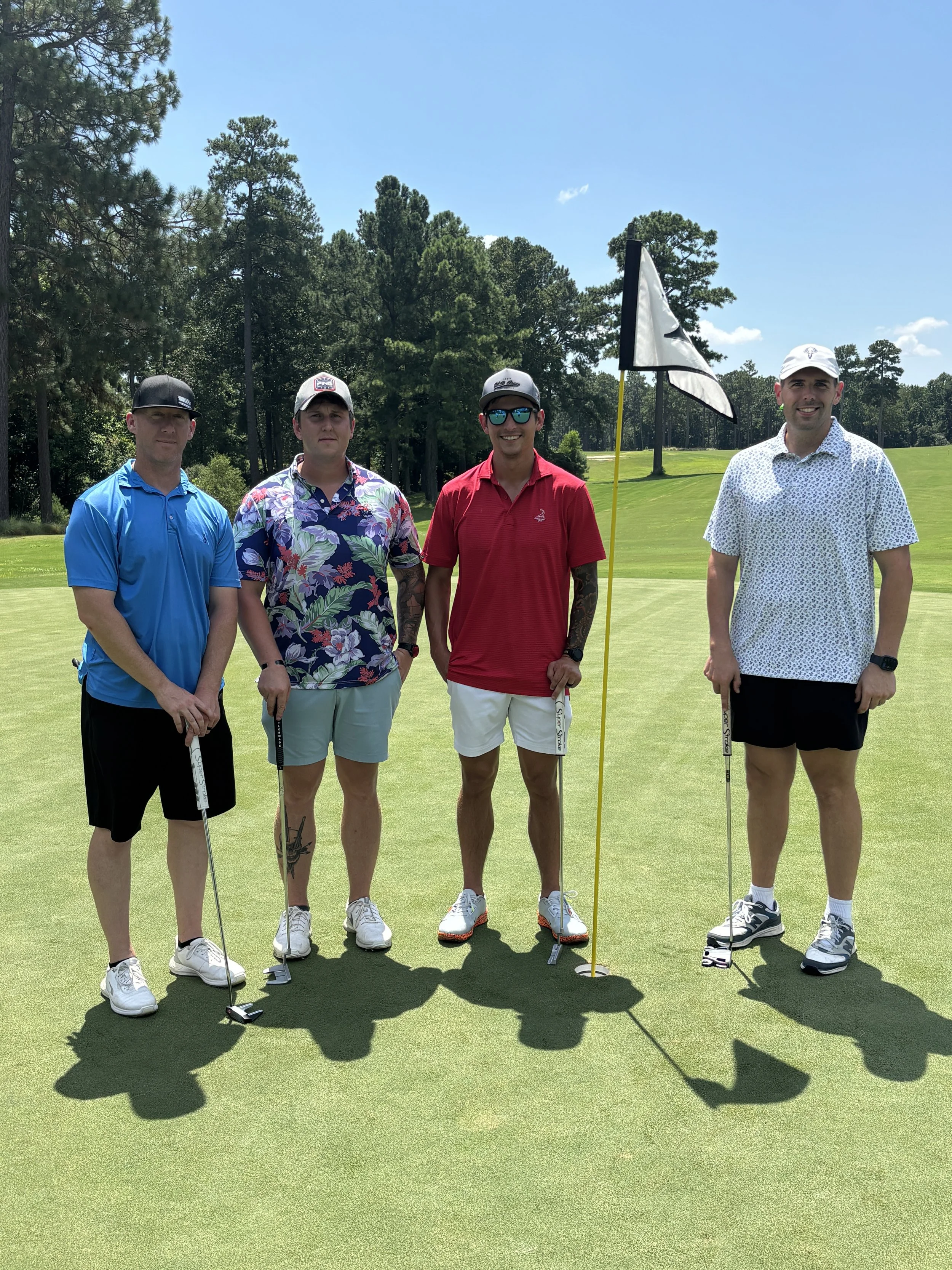 Four men standing on a golf course holding putters and a flagstick with a flag, under a blue sky with trees in the background.