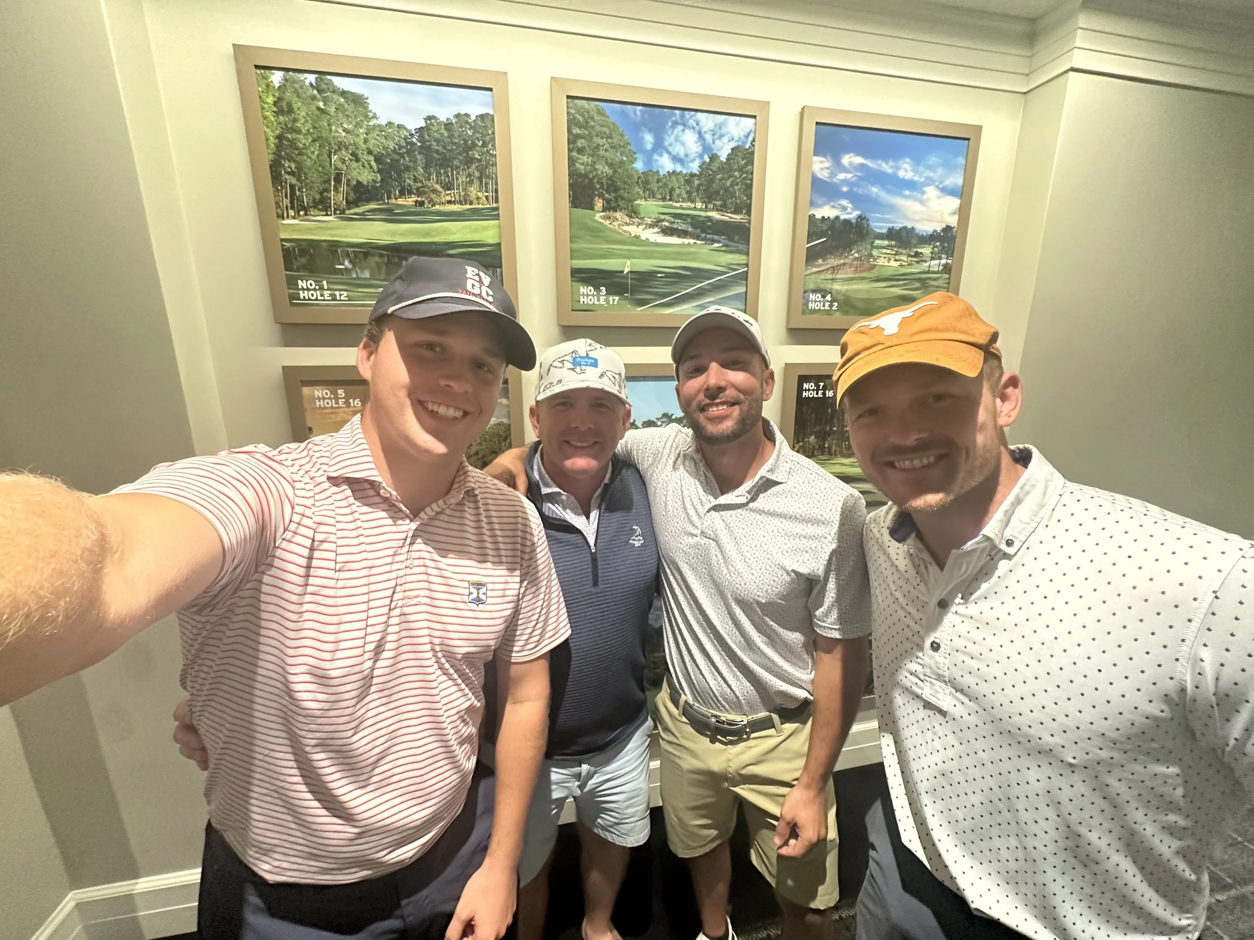 Four men taking a selfie in front of framed golf course photos, smiling, wearing golf caps and casual golf attire.