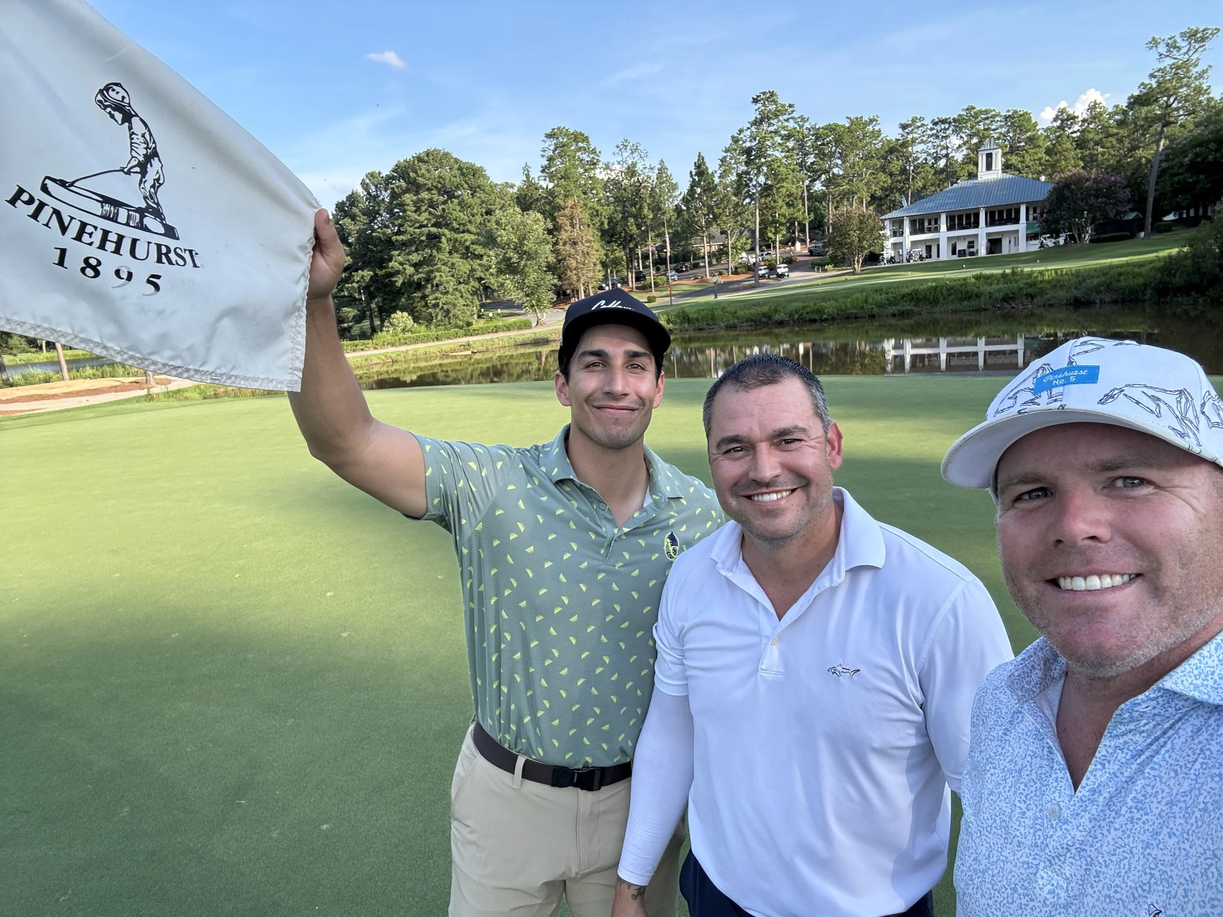 Three men taking a selfie on a golf course with trees, a pond, and a large white clubhouse in the background. One man holds a flag with a logo and the words 'Pinehurst 1895.'