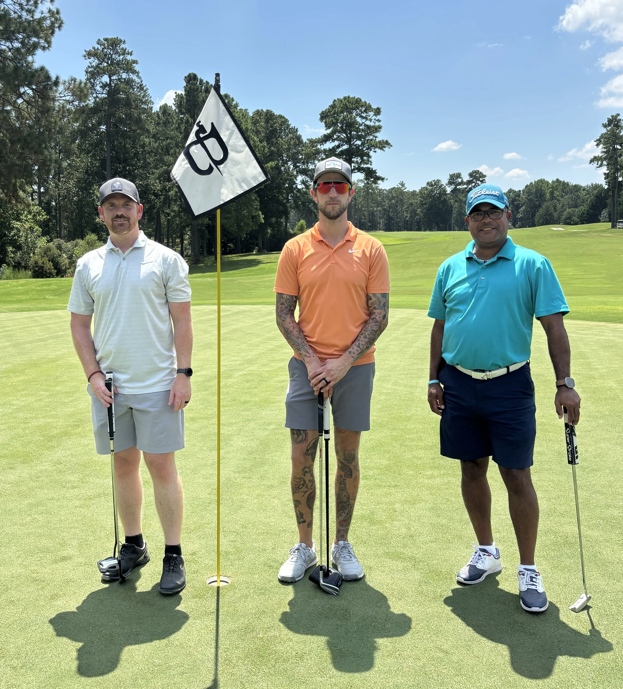 Three men standing on a golf course holding clubs, with a flag marking a hole behind them. They are dressed in golf attire, with trees and a blue sky in the background.