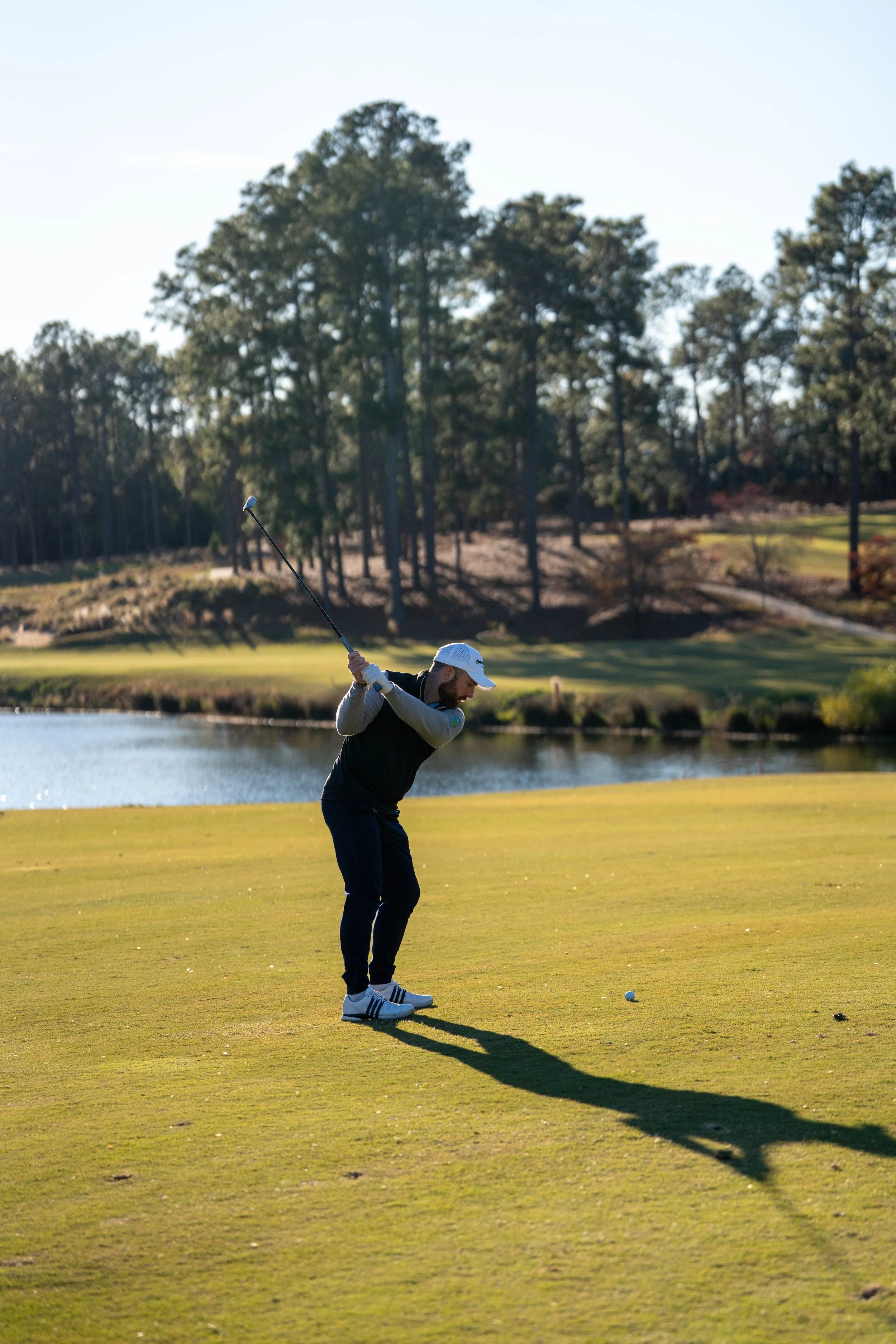 A man playing golf on a course near a pond, swinging his club with trees in the background during daylight.