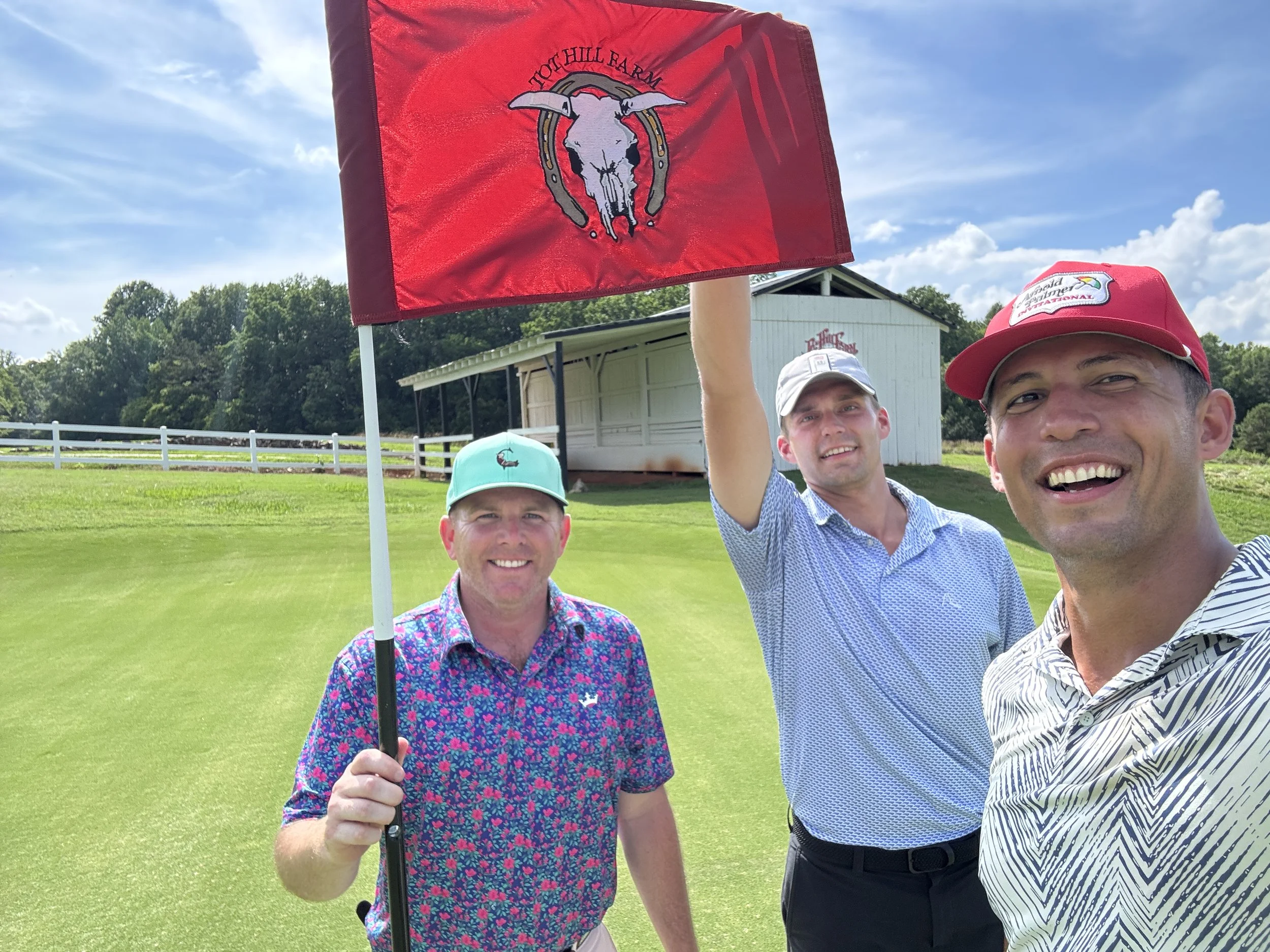 Three men on a golf course taking a selfie, one holding a red flag with a bull skull logo. The background features a green field, a white fence, and a small white structure, under a partly cloudy sky.