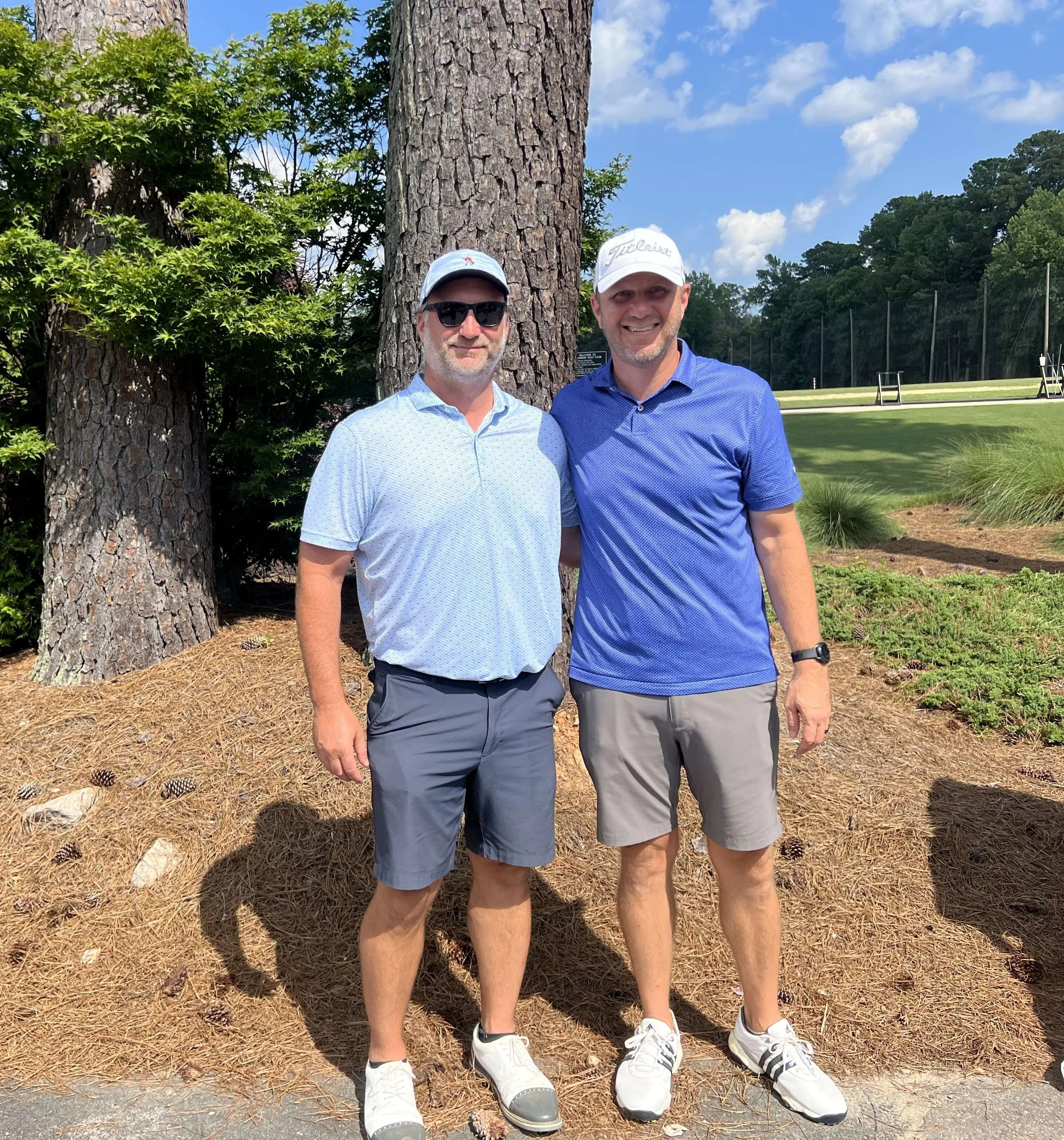 Two men in golf attire standing outdoors near large trees, smiling for a photo, with a grassy golf course and trees in the background.