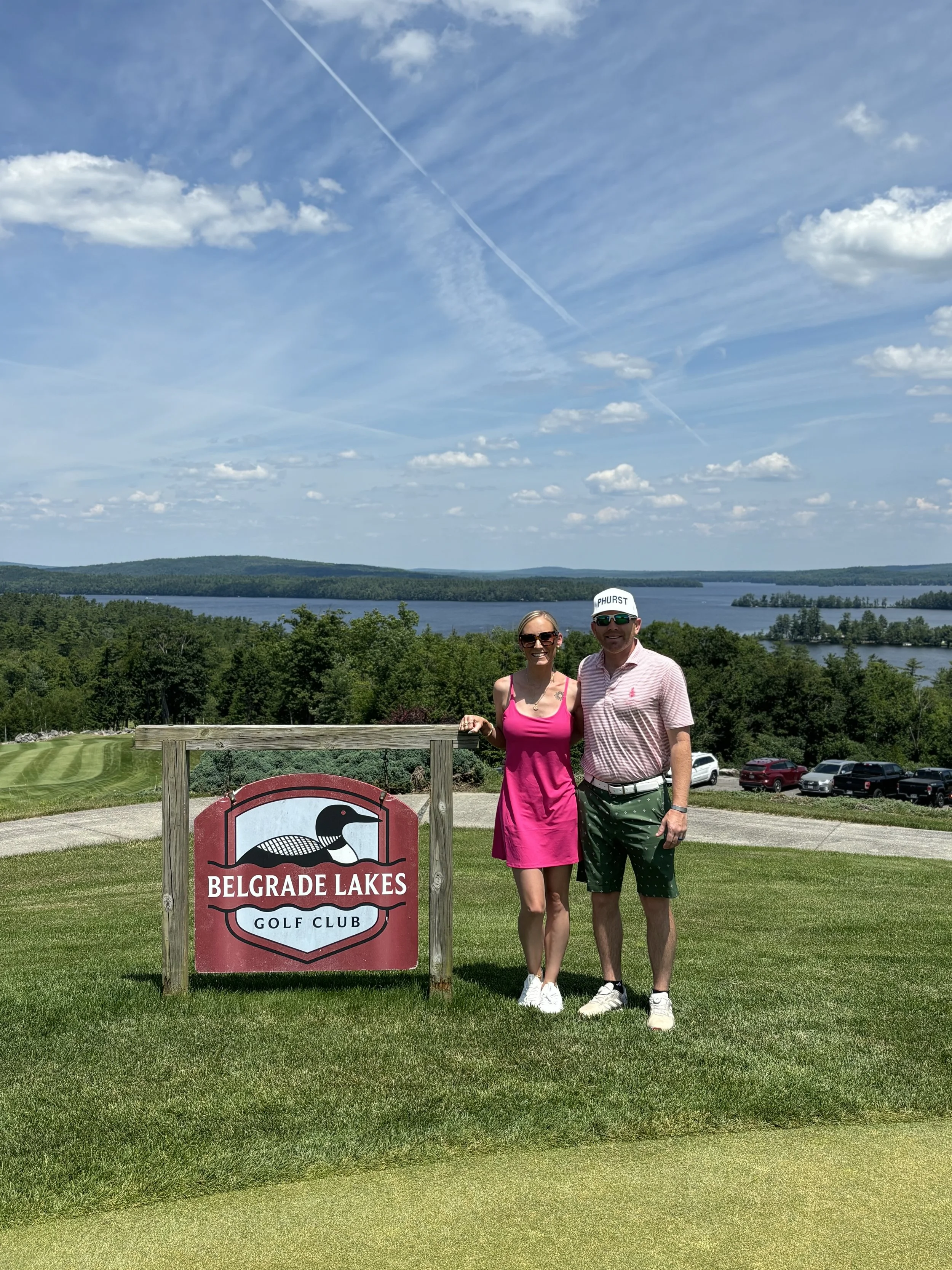 A couple standing outdoors at Belgrade Lakes Golf Club, with a lake and wooded area in the background, on a sunny day.