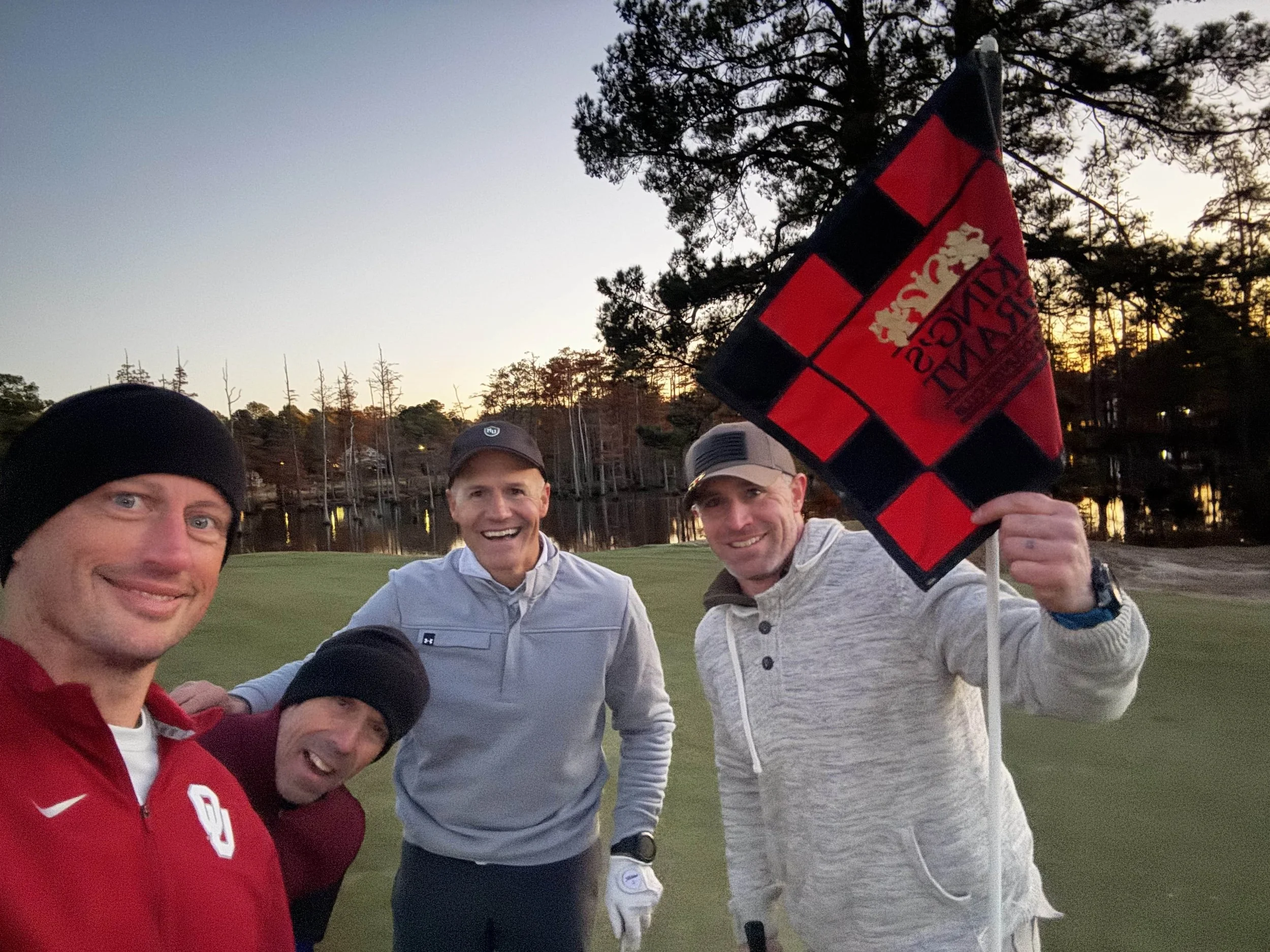 Four men smiling on a golf course during sunset, one holding a black and red checkered flag with a gold crest