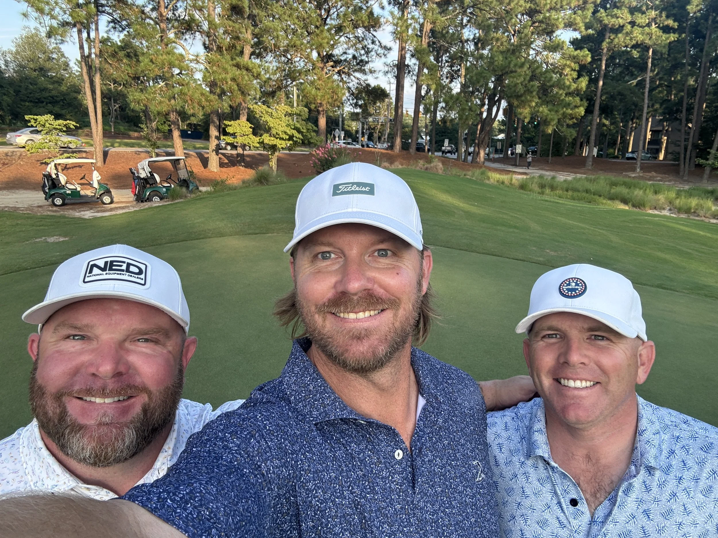 Three men taking a selfie on a golf course, wearing hats and casual shirts with golf carts and trees in the background.