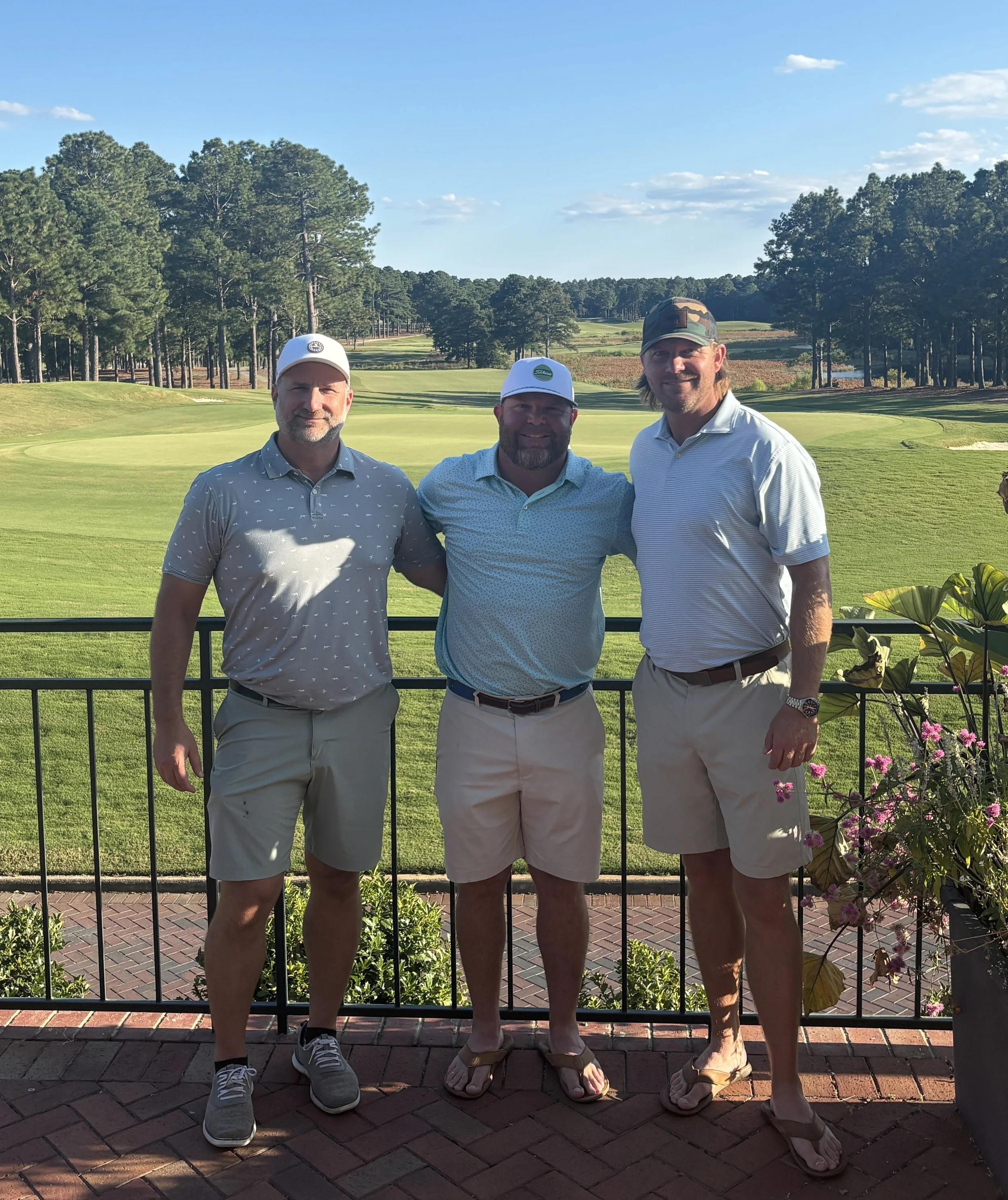 Three men standing on a balcony with a golf course in the background, wearing golf hats and casual summer clothing.