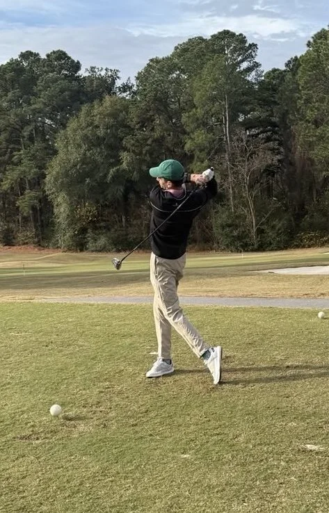 A person in a black jacket, beige pants, and white shoes swinging a golf club on a golf course, with a golf ball in front of them. Trees and a cloudy sky are in the background.