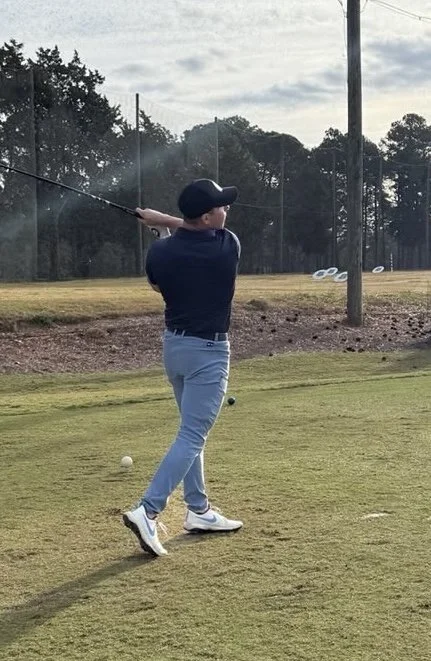 A person in a navy polo shirt, light blue jeans, and white shoes, swinging a golf club on a golf course during daytime.