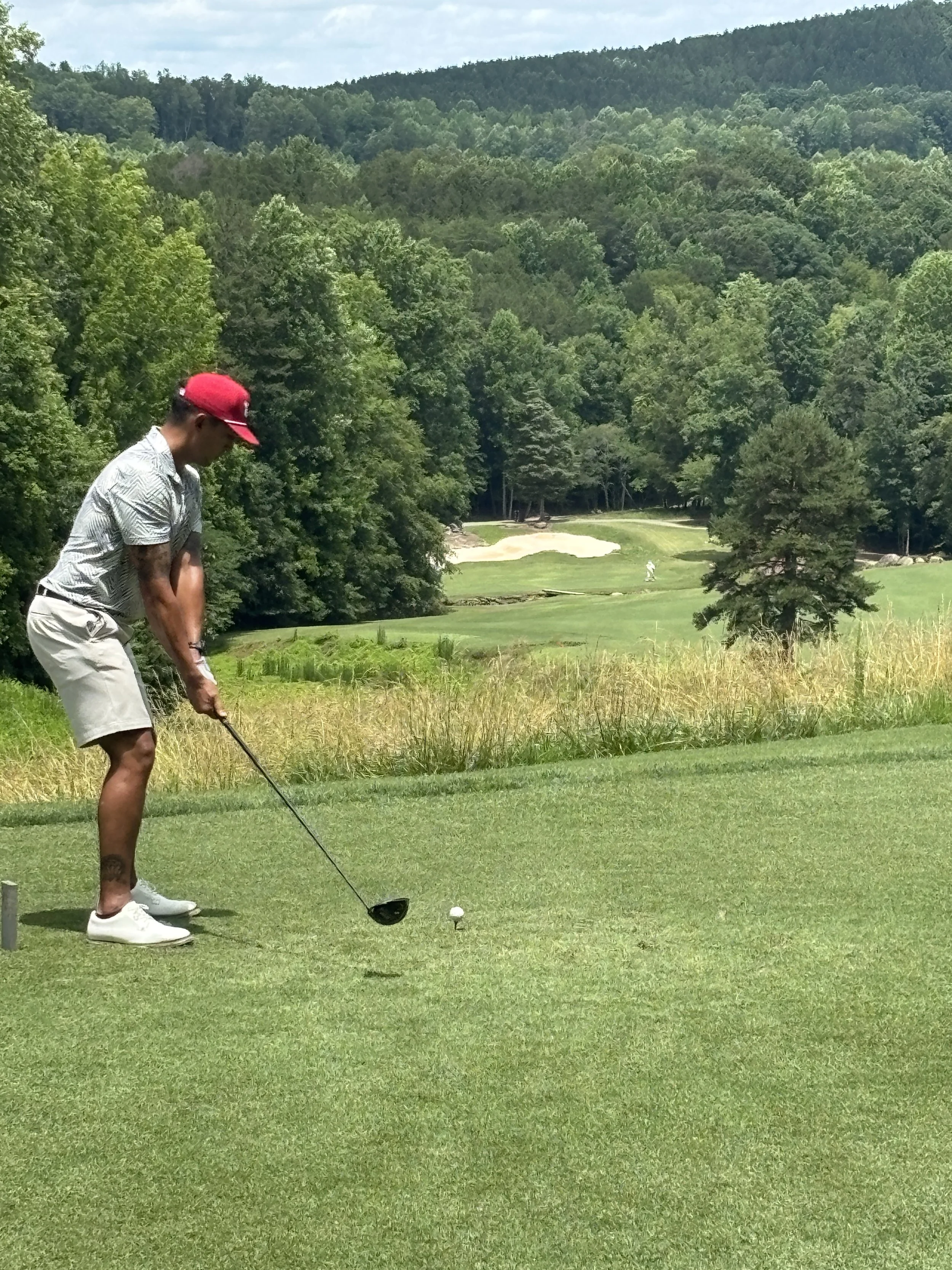 Man in a red cap tees off on a golf course surrounded by green trees and hills.