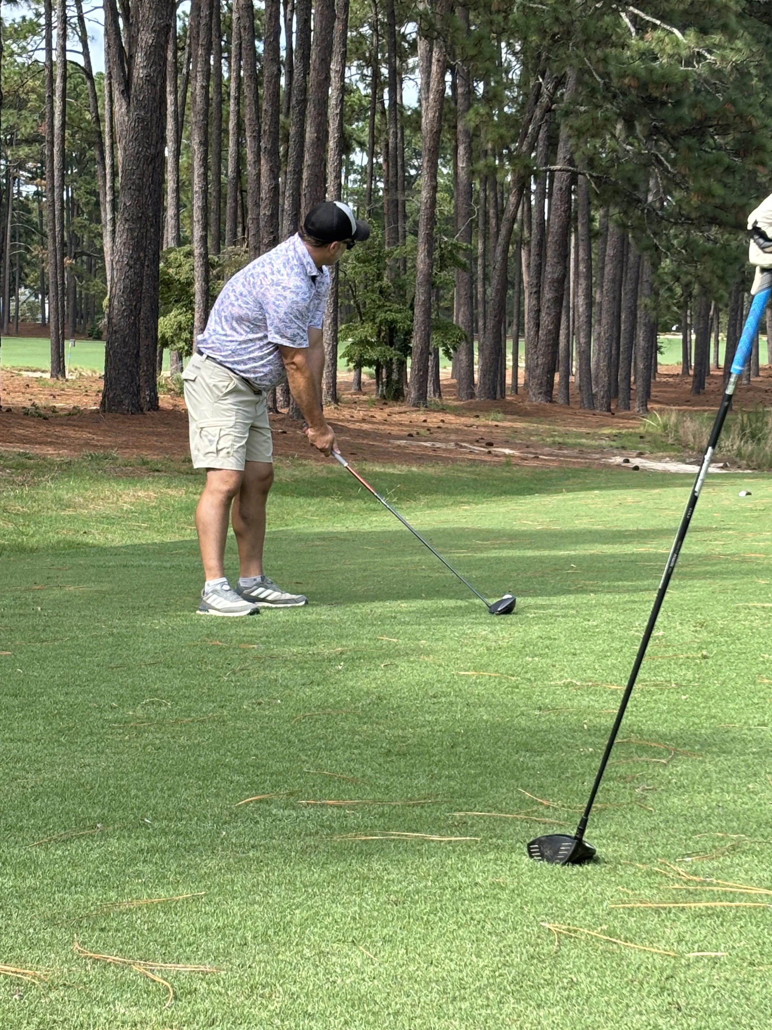 A man in a floral shirt, shorts, and a cap preparing to tee off on a golf course, with trees in the background and two golf clubs on the grass.