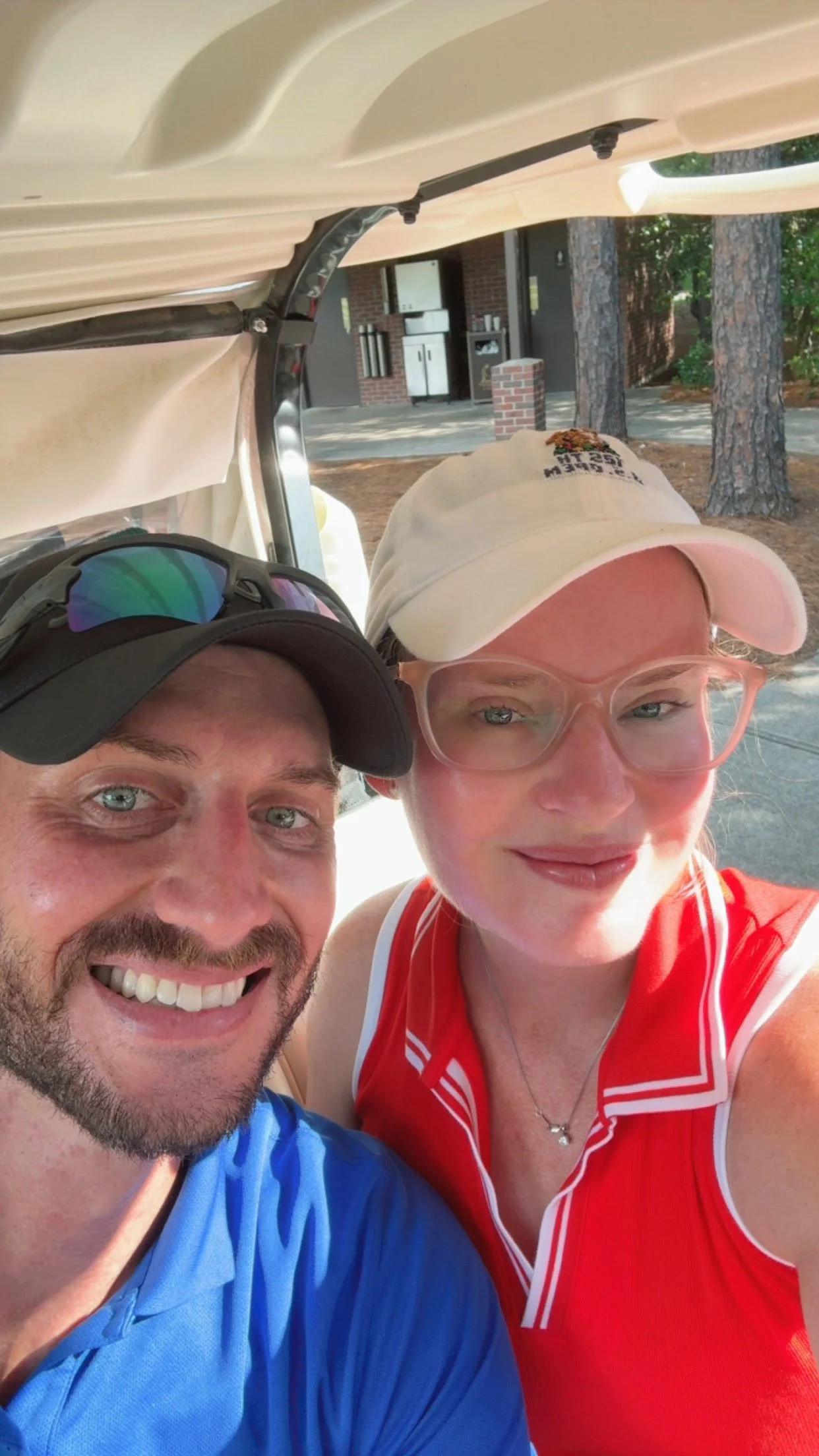 Two smiling people taking a selfie outdoors near a golf cart, with trees and a brick building in the background.