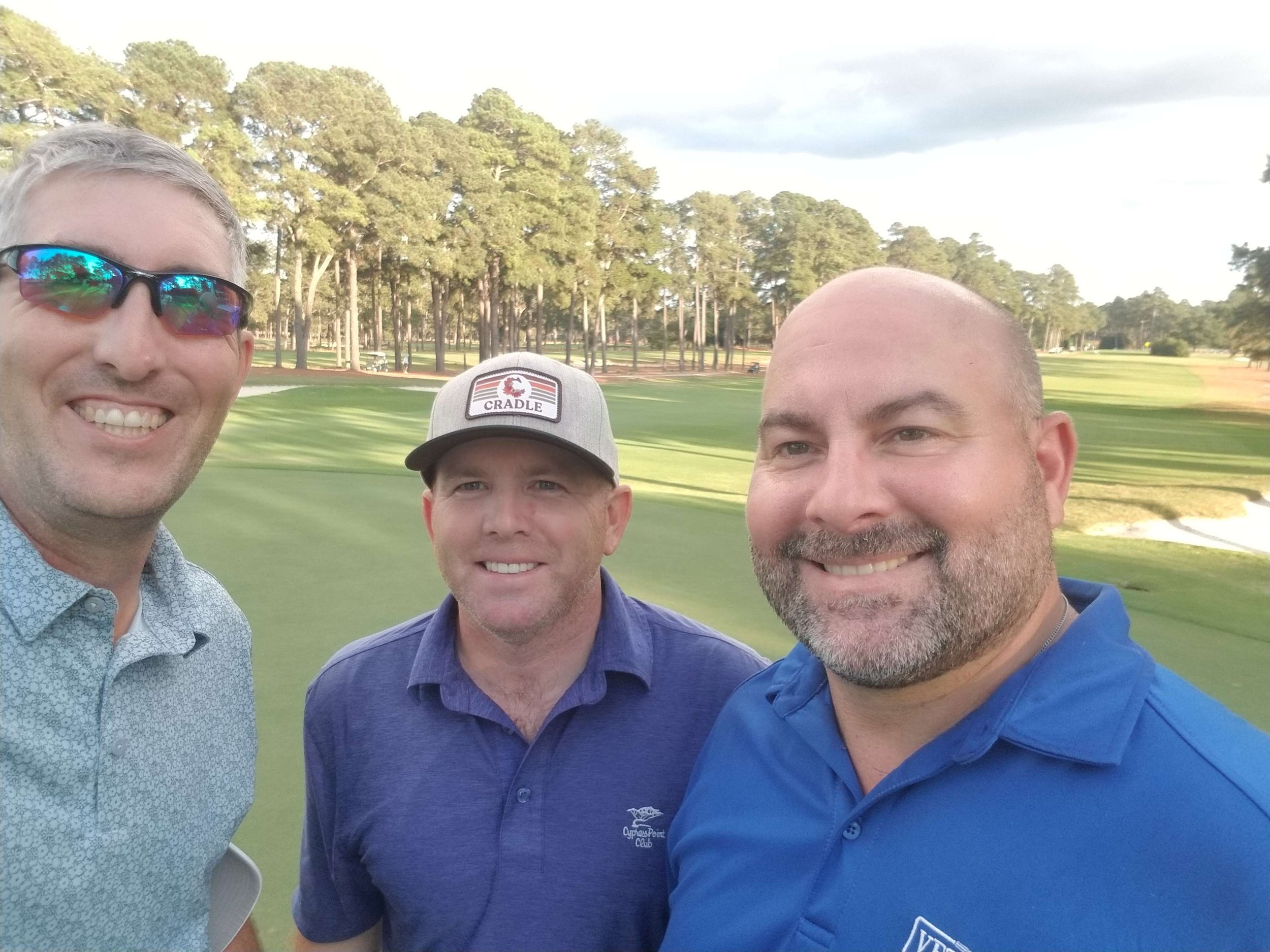 Three men taking a selfie on a golf course with green grass, trees, and a partly cloudy sky in the background.
