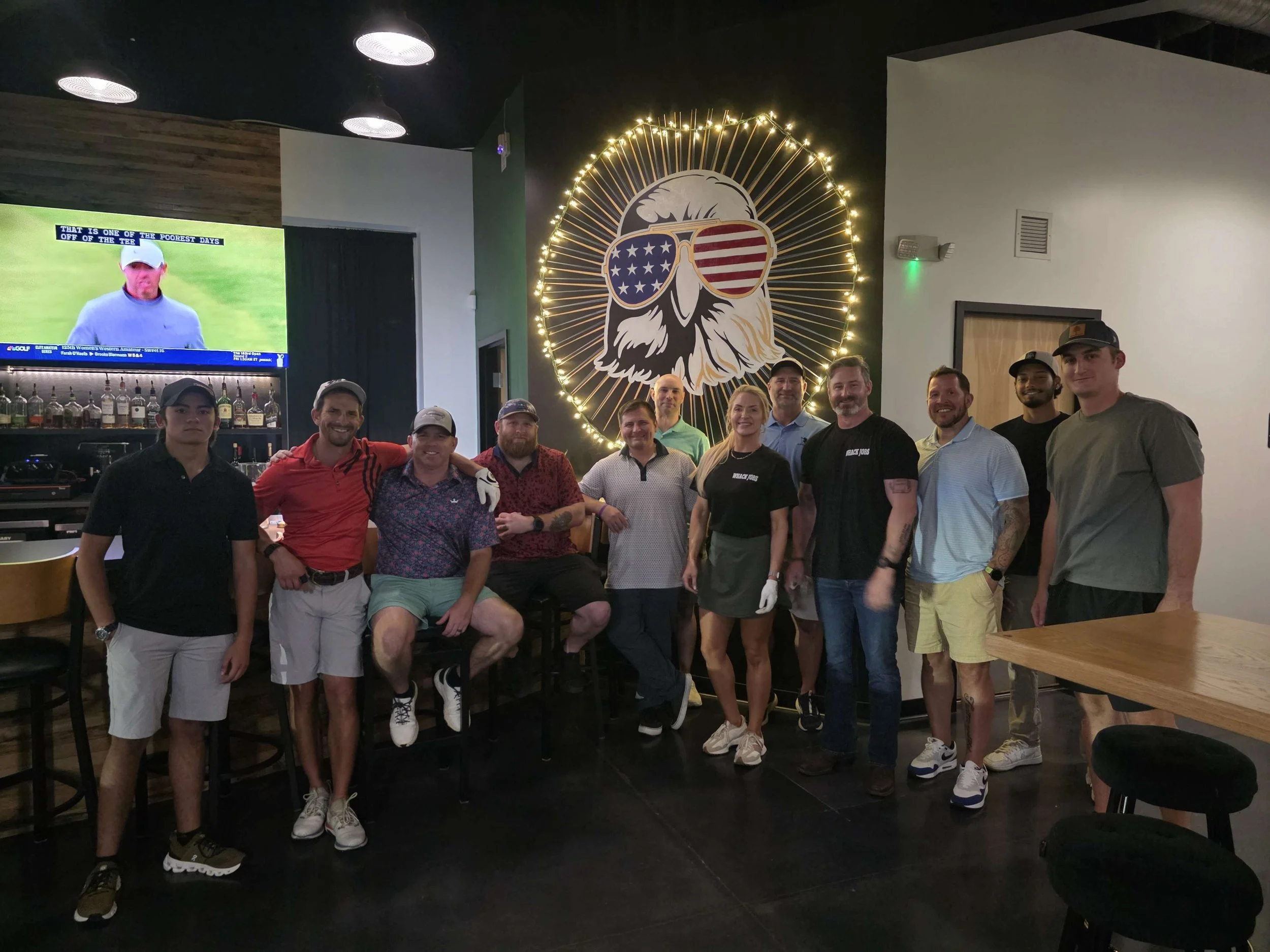 Group of twelve people posing inside a bar with a large eagle mural wearing sunglasses decorated with American flag patterns."