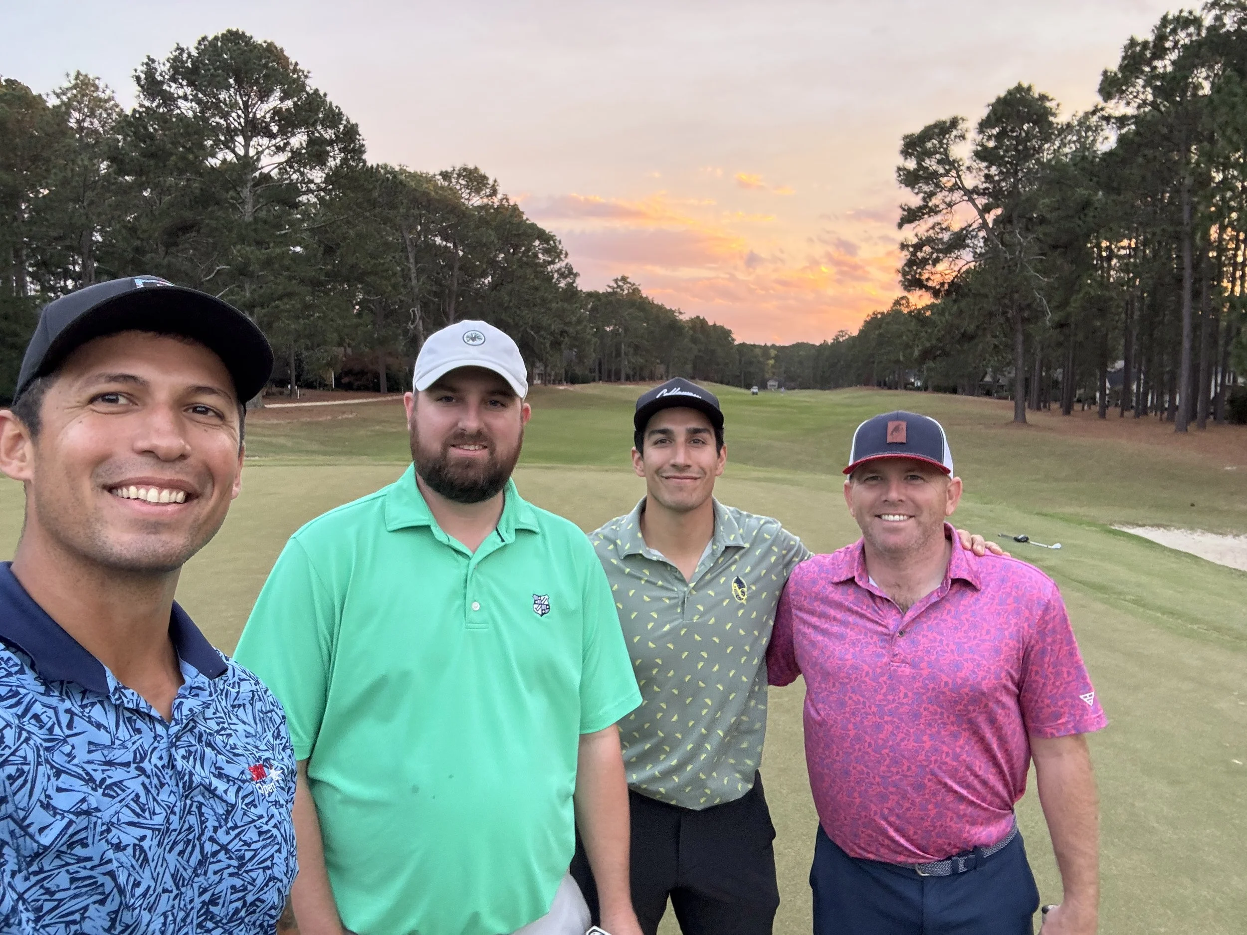 Four men smiling on a golf course during sunset, dressed in golf shirts and caps.
