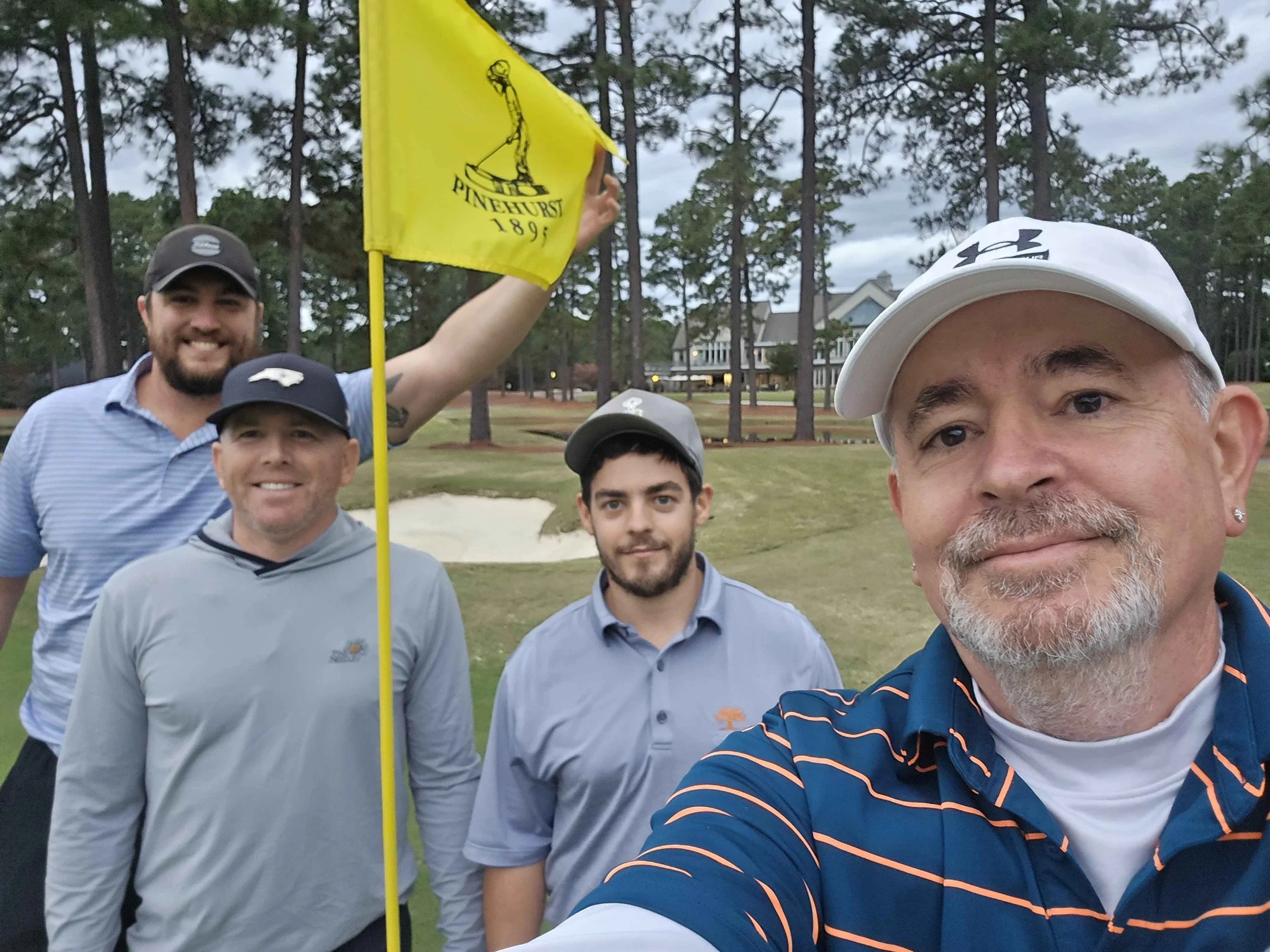 Four men at a golf course, smiling for a selfie, with a yellow flag and golf course in the background.