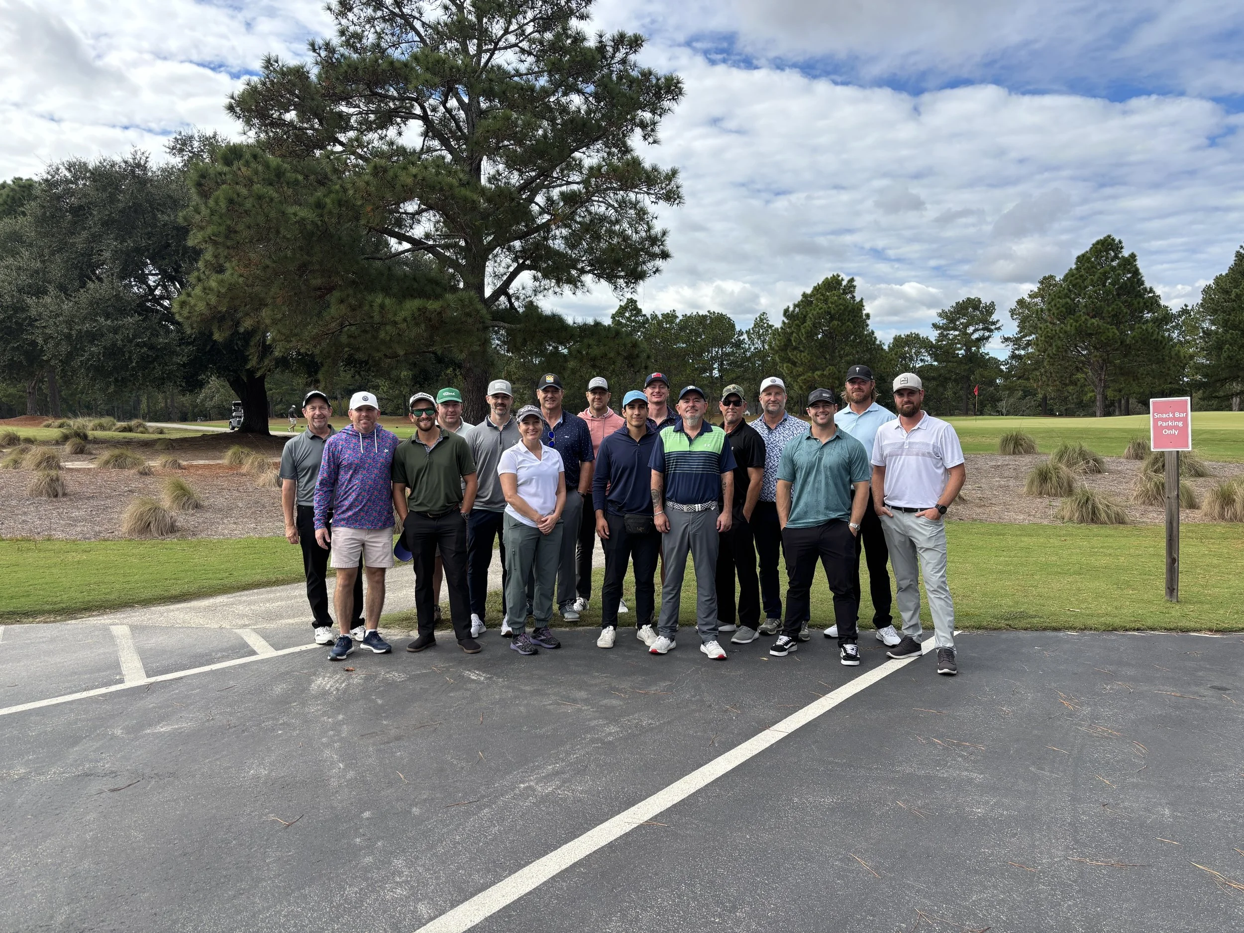 Group of 15 people standing on a golf course, smiling, with trees and a cloudy sky in the background.