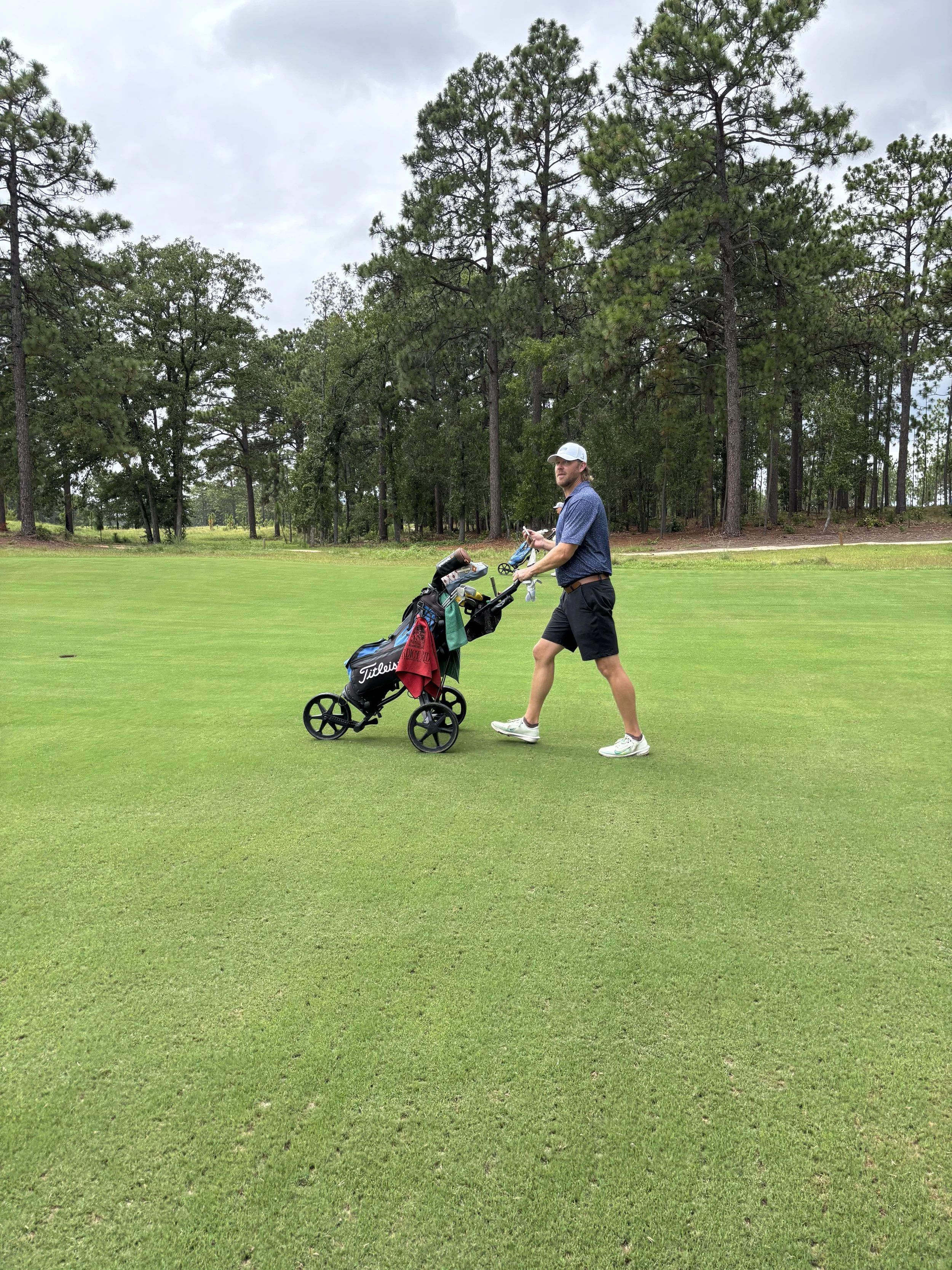 A man standing on a golf course next to his golf cart, holding golf clubs and looking at something in his hand, with trees in the background.