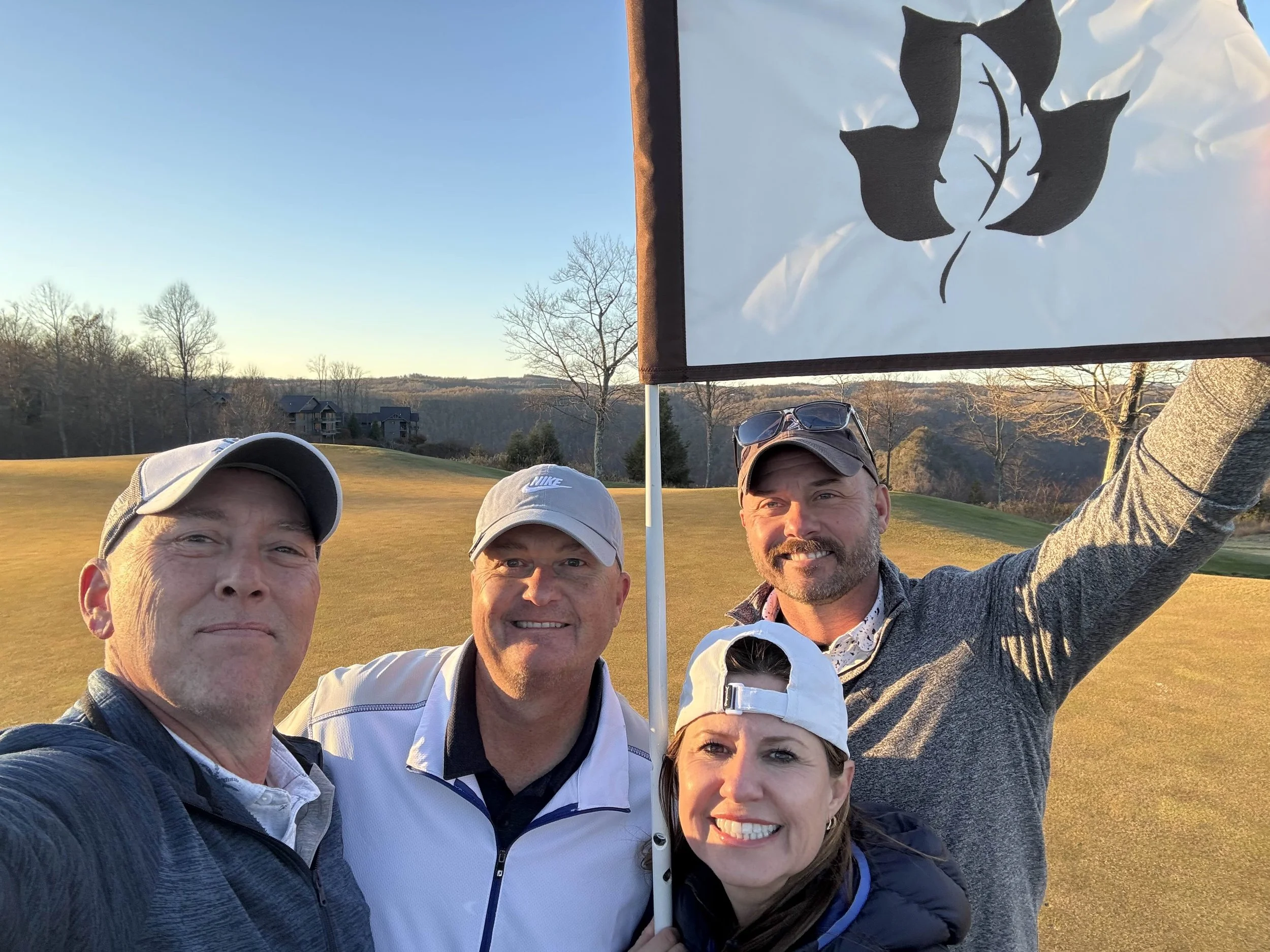 Four people on a golf course taking a selfie with a flag. The flag has a black leaf design. The background shows a clear sky, leafless trees, and houses in the distance.