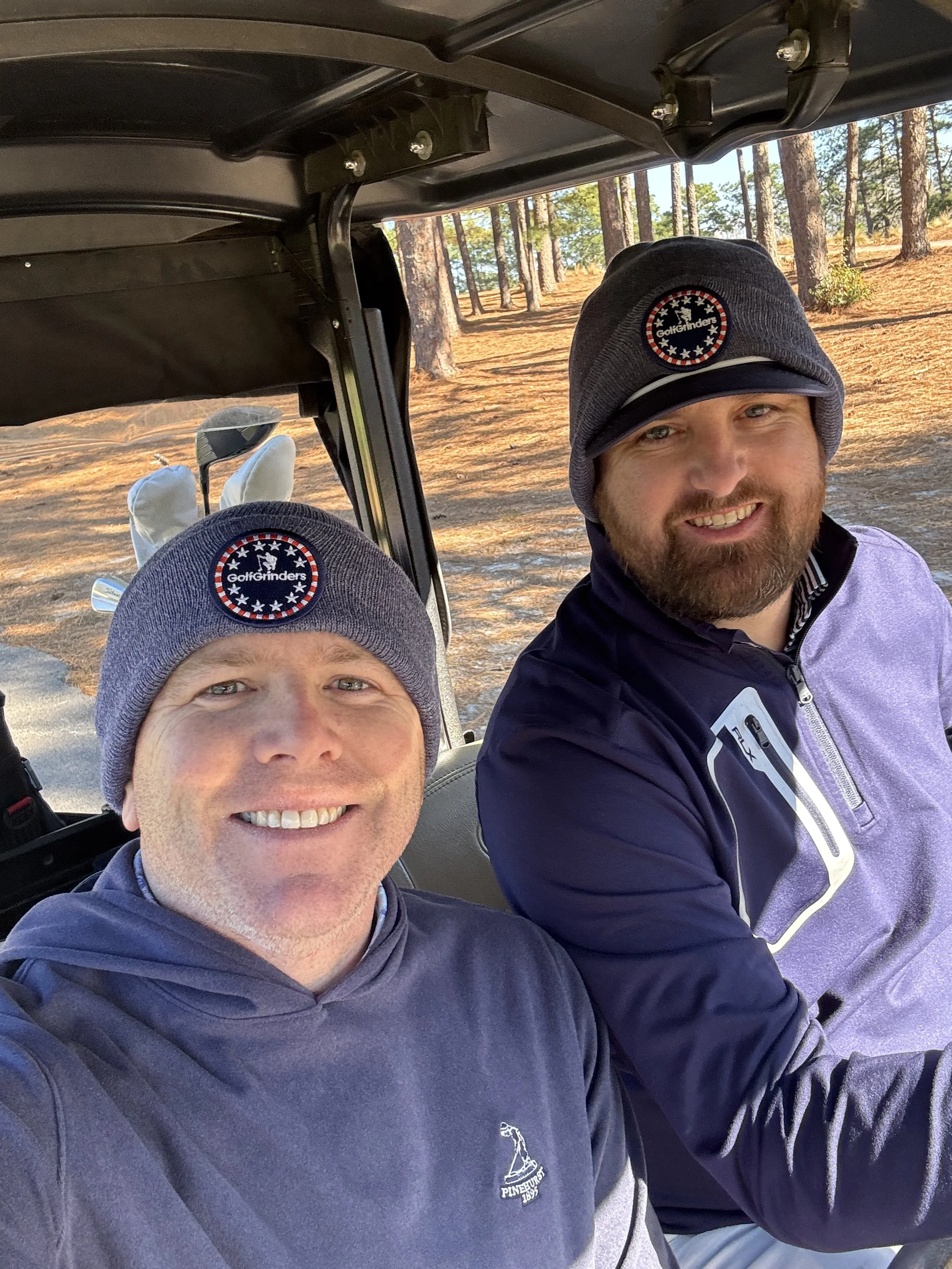 Two men wearing baseball caps and jackets taking a selfie on a golf course, with trees and sunlight in the background.