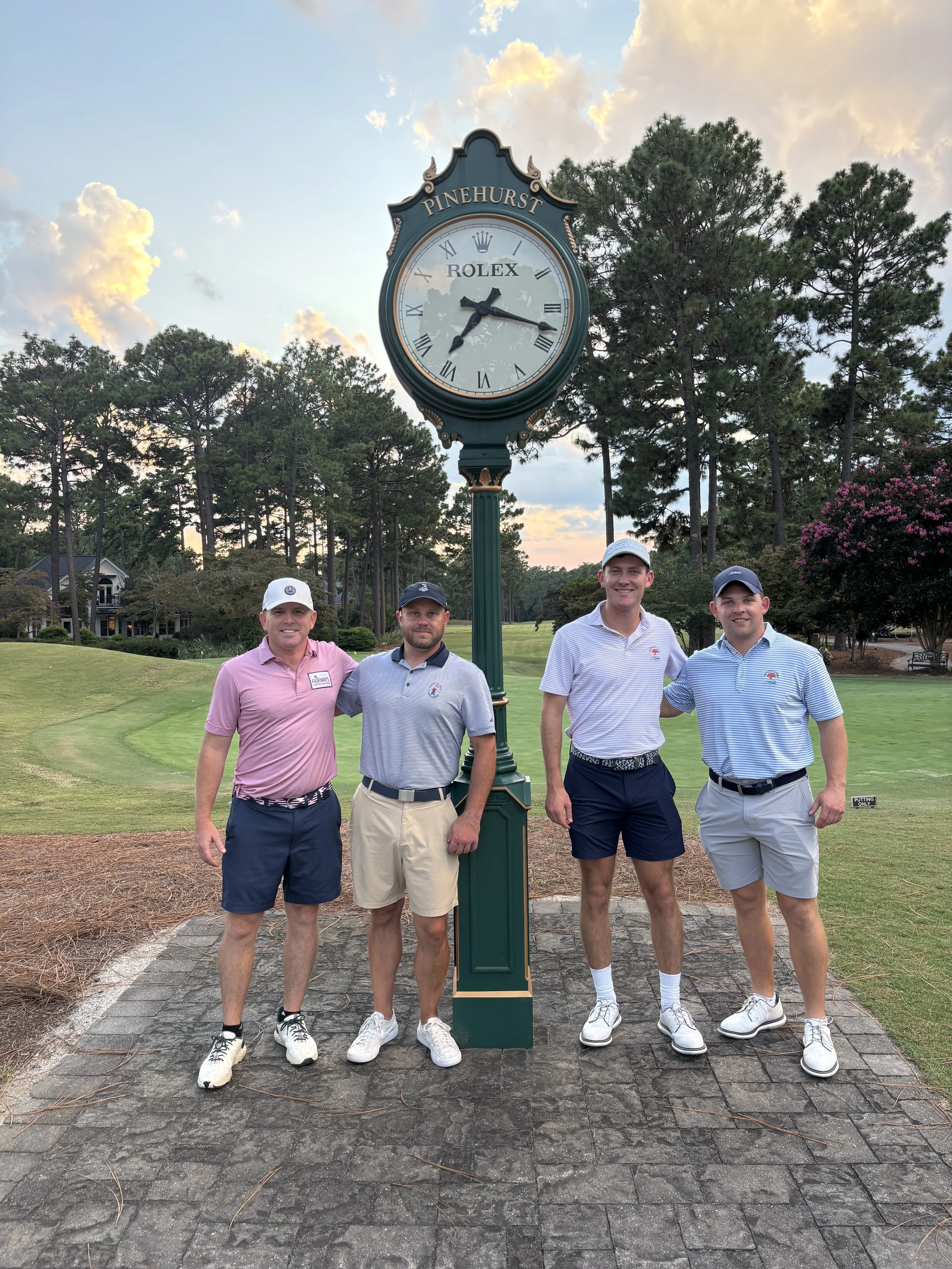 Four men standing outdoors on a golf course near a tall clock with the words 'Pinehurst' and 'Rolex' on it. They are dressed in golf attire and smiling at the camera, with trees and a partly cloudy sky in the background.