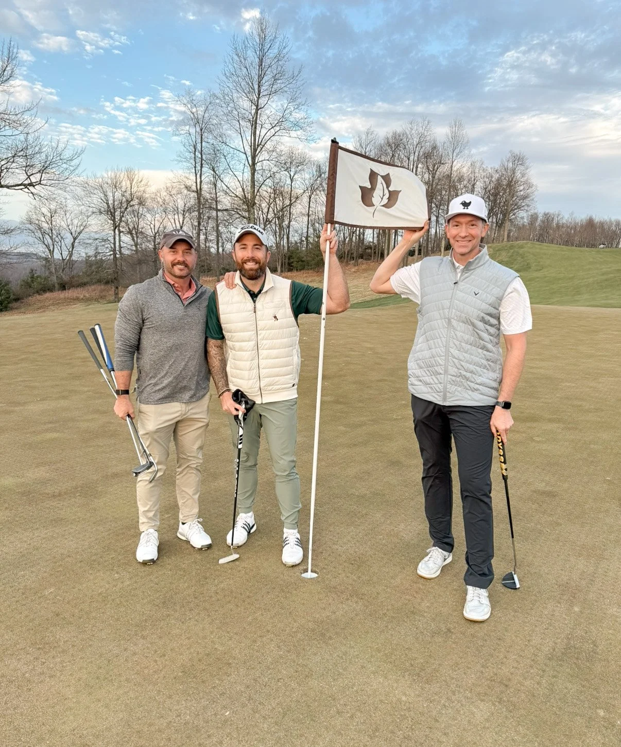Four men on a golf course with trees in the background. Two of them are holding golf clubs, and one is holding a flag with a leaf logo. All are smiling and dressed in golf attire.