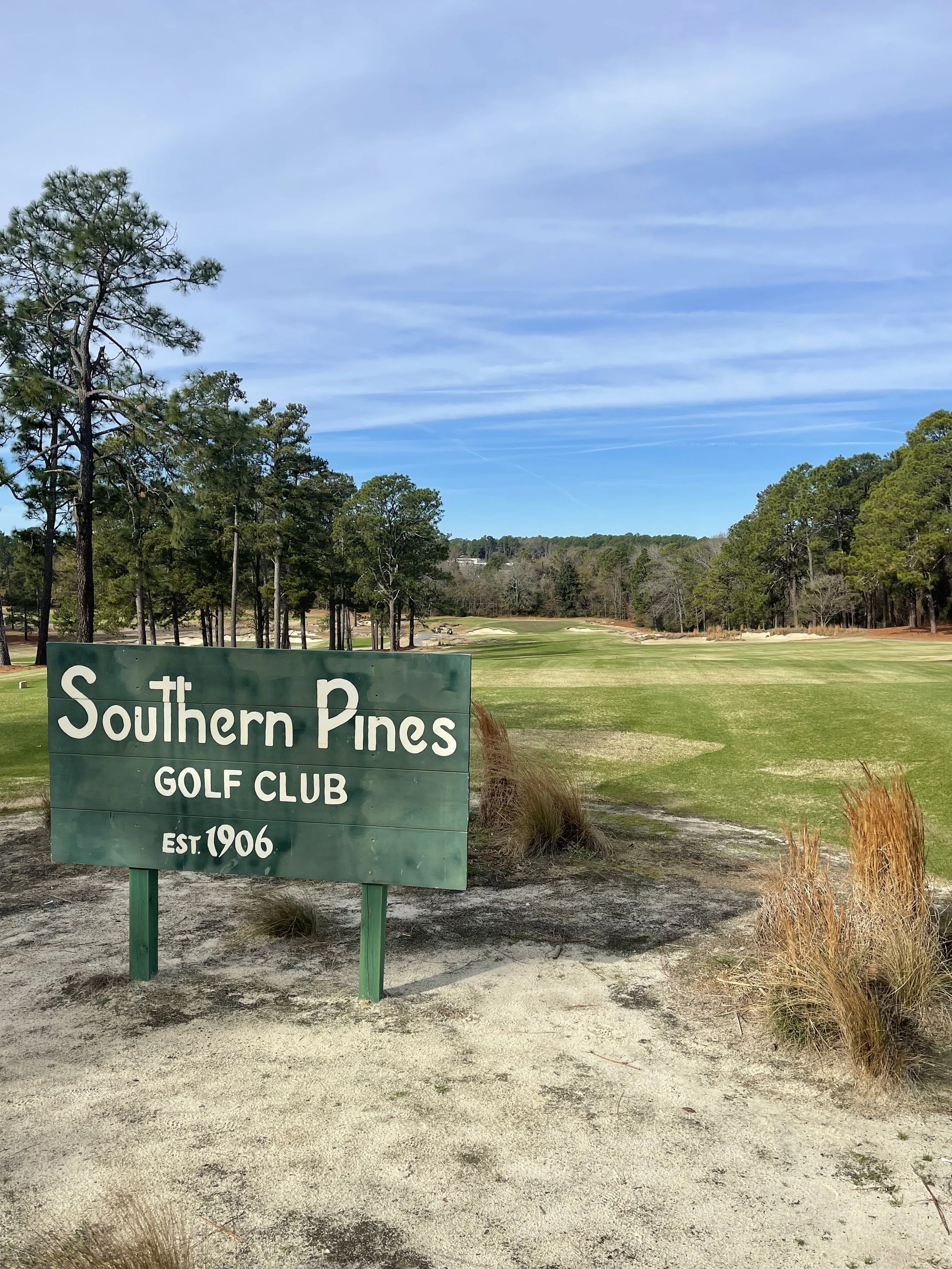 Sign for Southern Pines Golf Club established in 1906 at the entrance, with a view of the golf course and trees in the background.