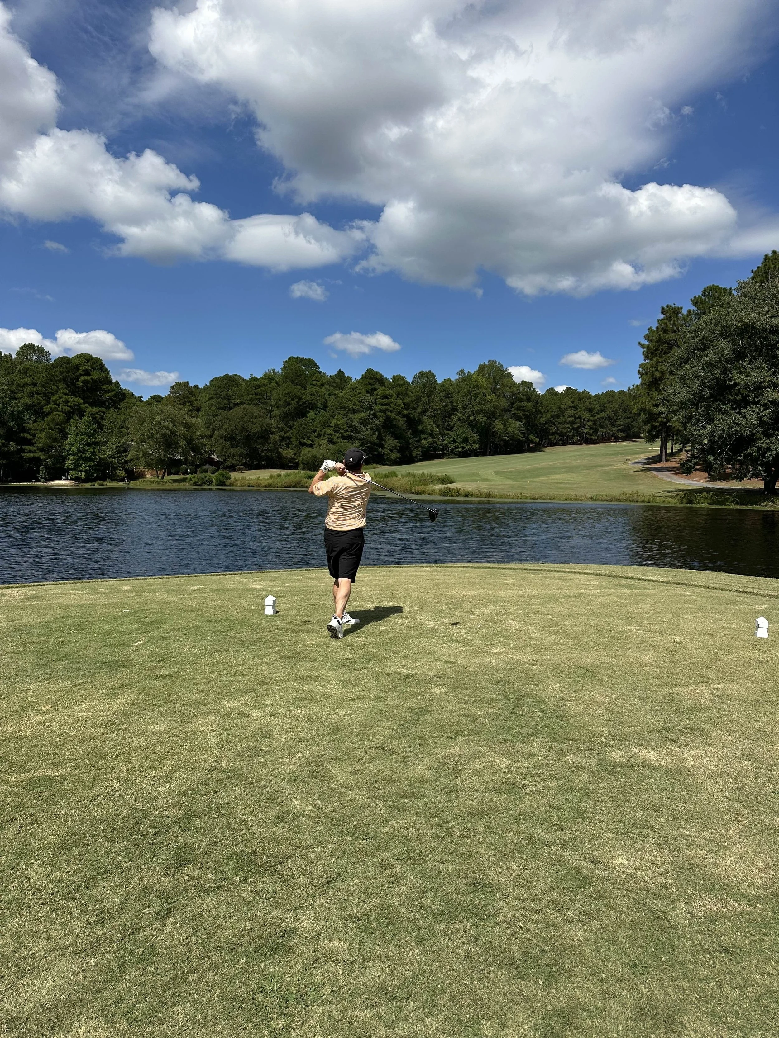A person playing golf on a course near a pond, with trees and a partly cloudy sky in the background.