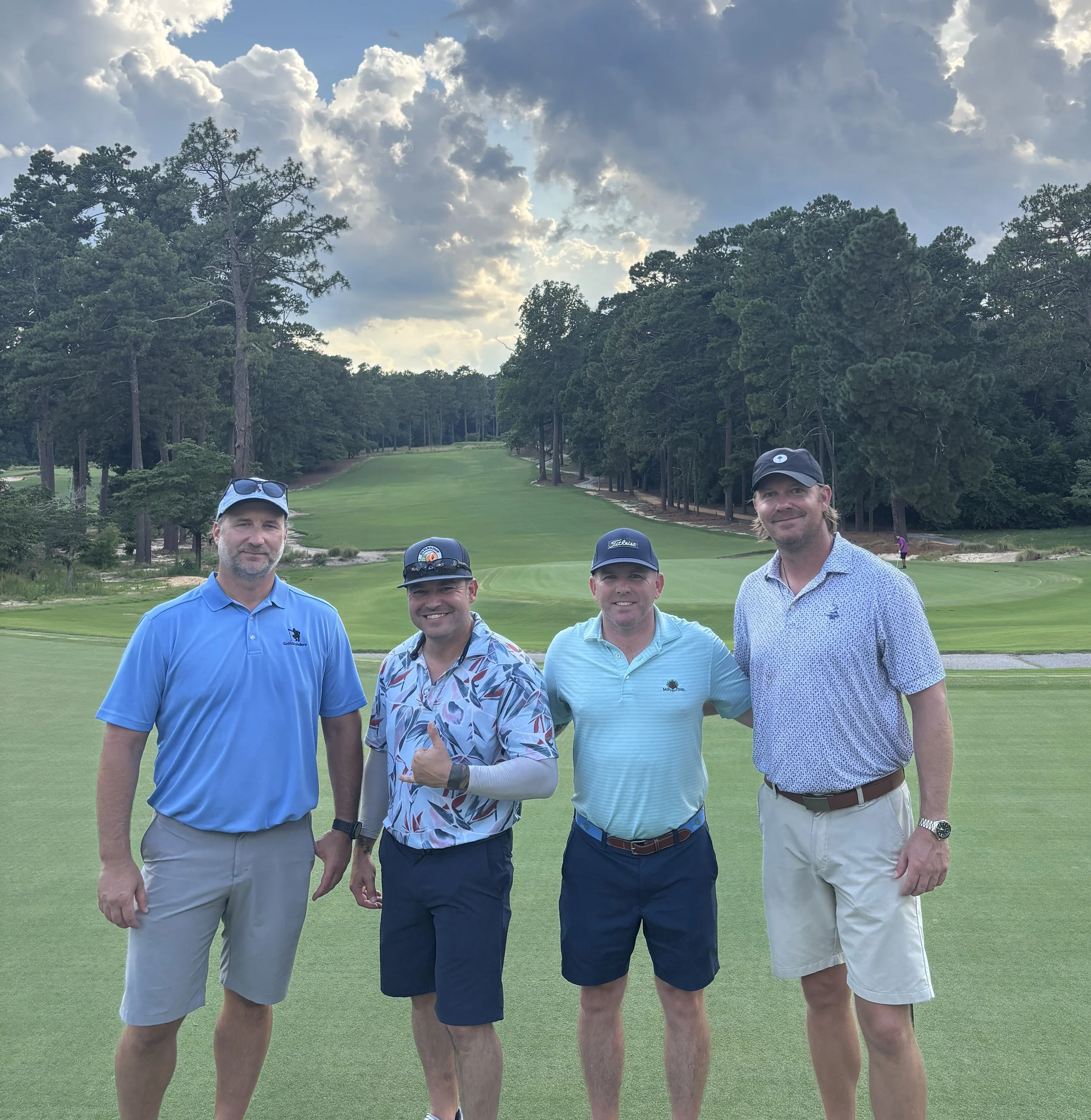 Four men standing together on a golf course, smiling at the camera, with trees and a cloudy sky in the background.