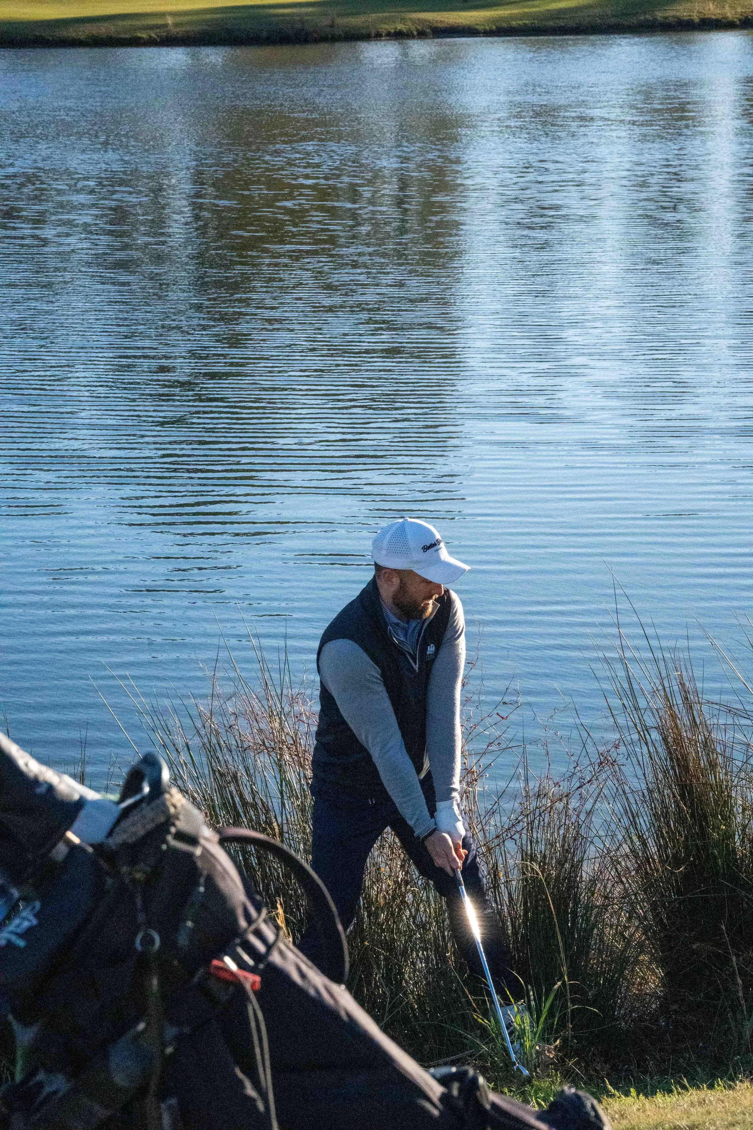 A man in a white cap and dark vest stands near a lake's edge, preparing to swing a golf club amidst tall grass.