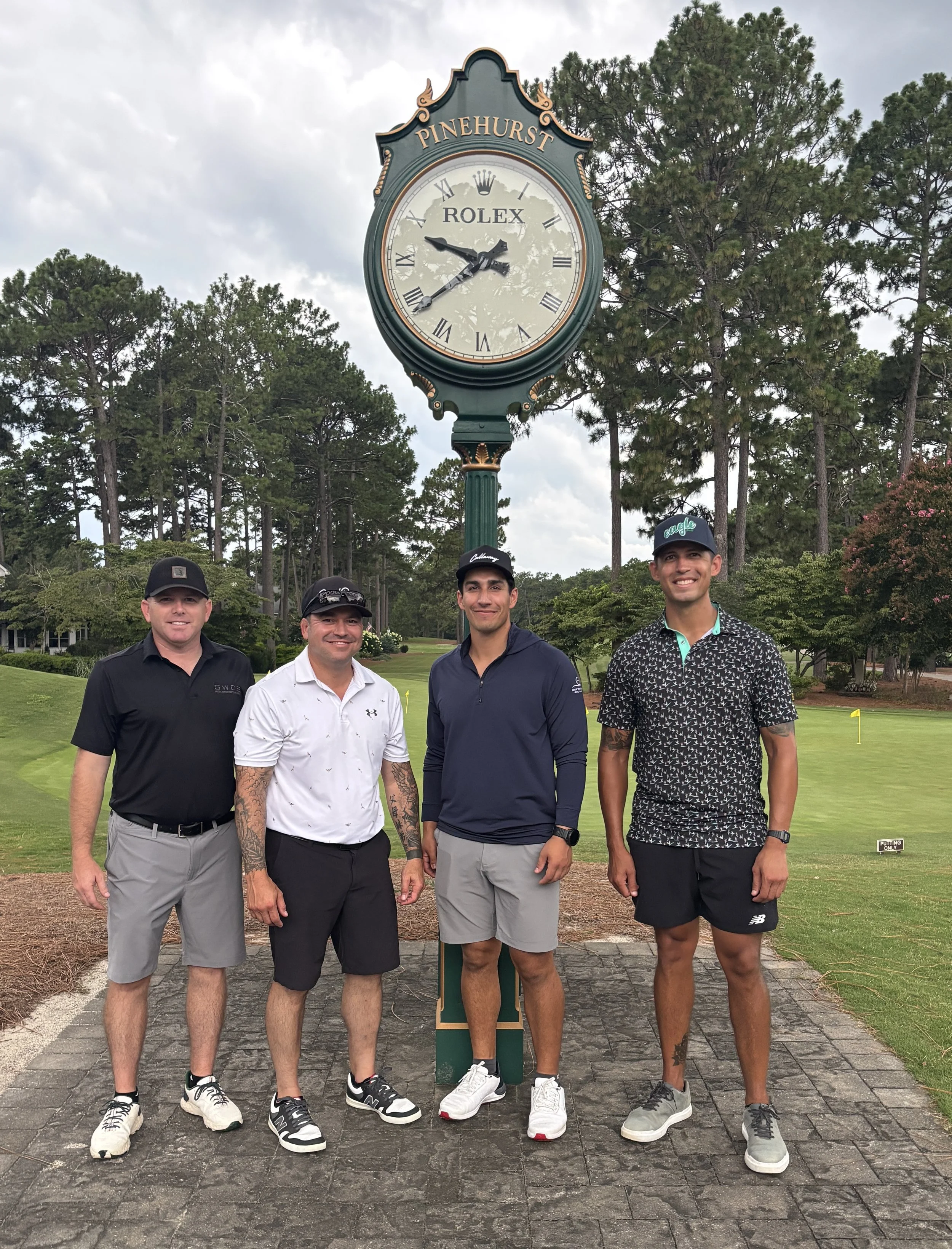 Four men standing in front of a large clock on a pole at Pinehurst golf course, surrounded by trees and a cloudy sky.