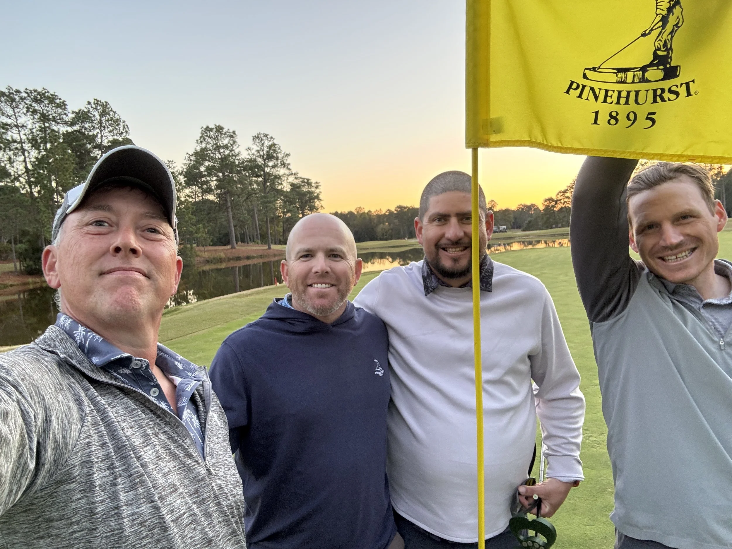 Four men on a golf course at sunset, taking a selfie with a yellow Pinehurst flag. They are smiling, dressed in golf attire, with trees and water in the background.