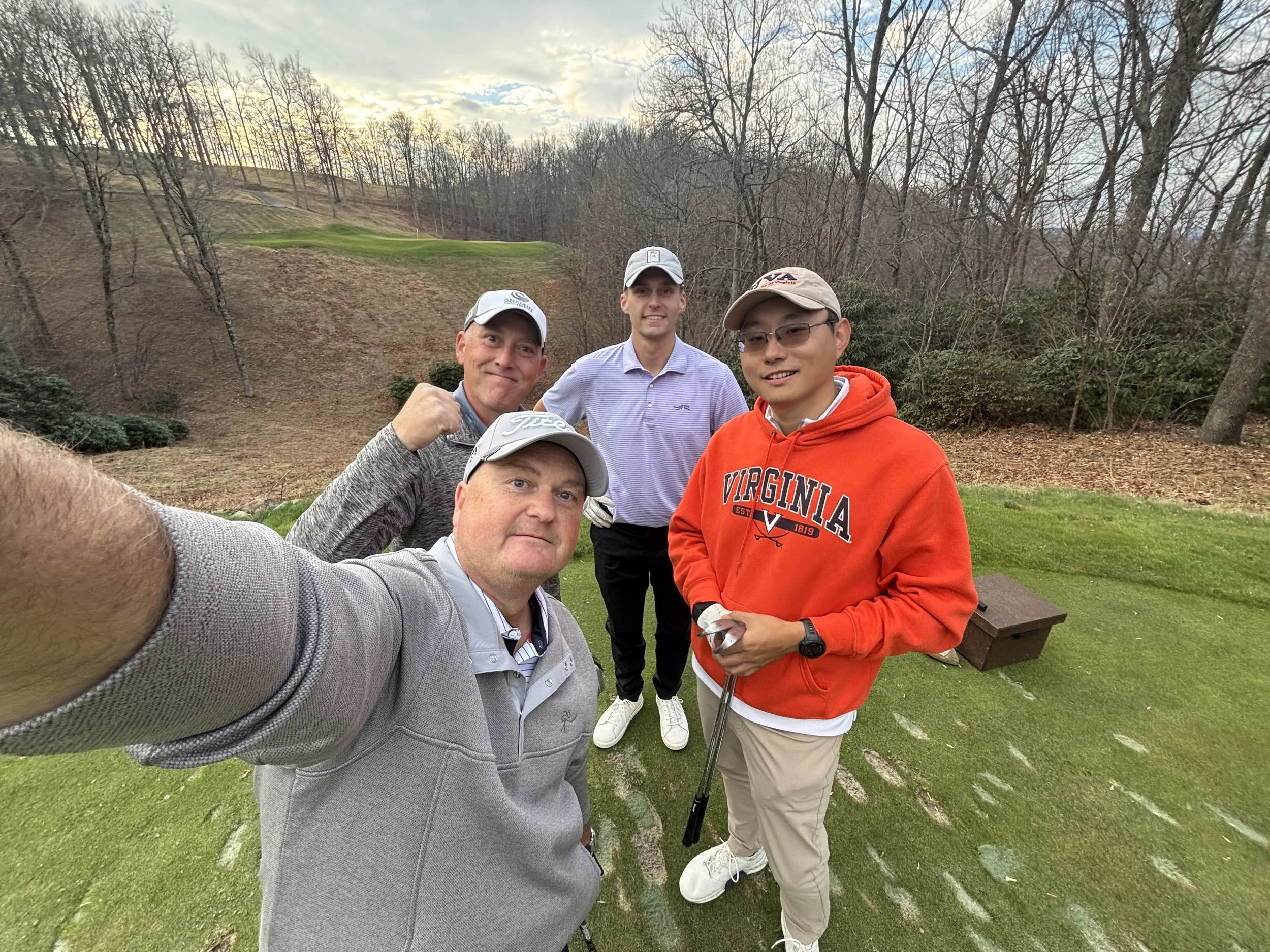 Group of five men on a golf course taking a selfie. One man in front holding the camera, wearing a gray pullover, and the others in the background, wearing golf caps and casual shirts or hoodies. They are standing on a grassy area with a hill and tre