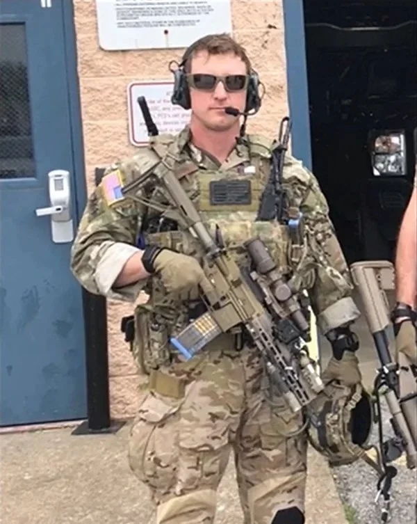 A person dressed in military camouflage gear with tactical equipment, holding a firearm, wearing sunglasses, and a headset with a microphone. The background shows a blue door and a beige wall.