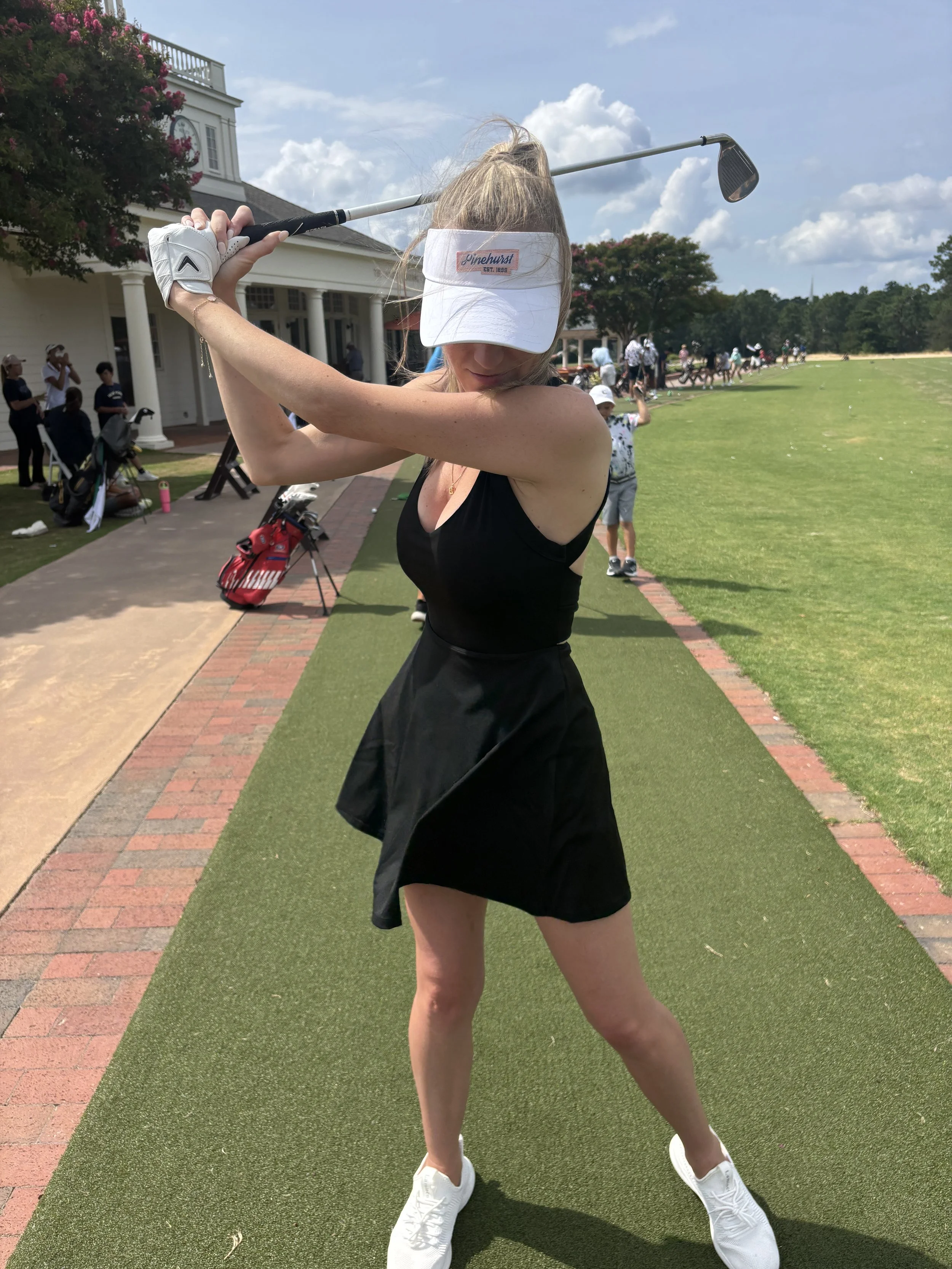 A woman in a black sleeveless dress and white sneakers swings a golf club, preparing to hit a golf ball at a driving range on a sunny day.
