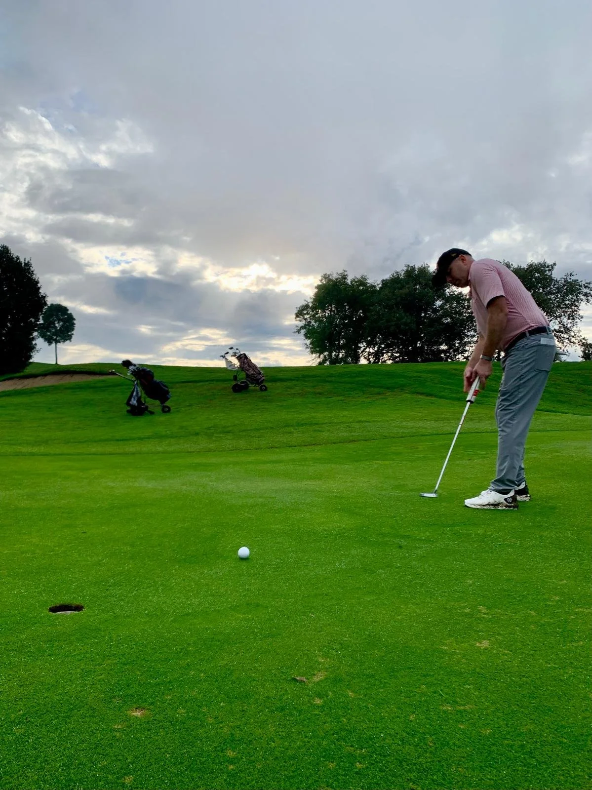 A golfer preparing to putt on a golf course with golf bags and carts on the green, under a partly cloudy sky.