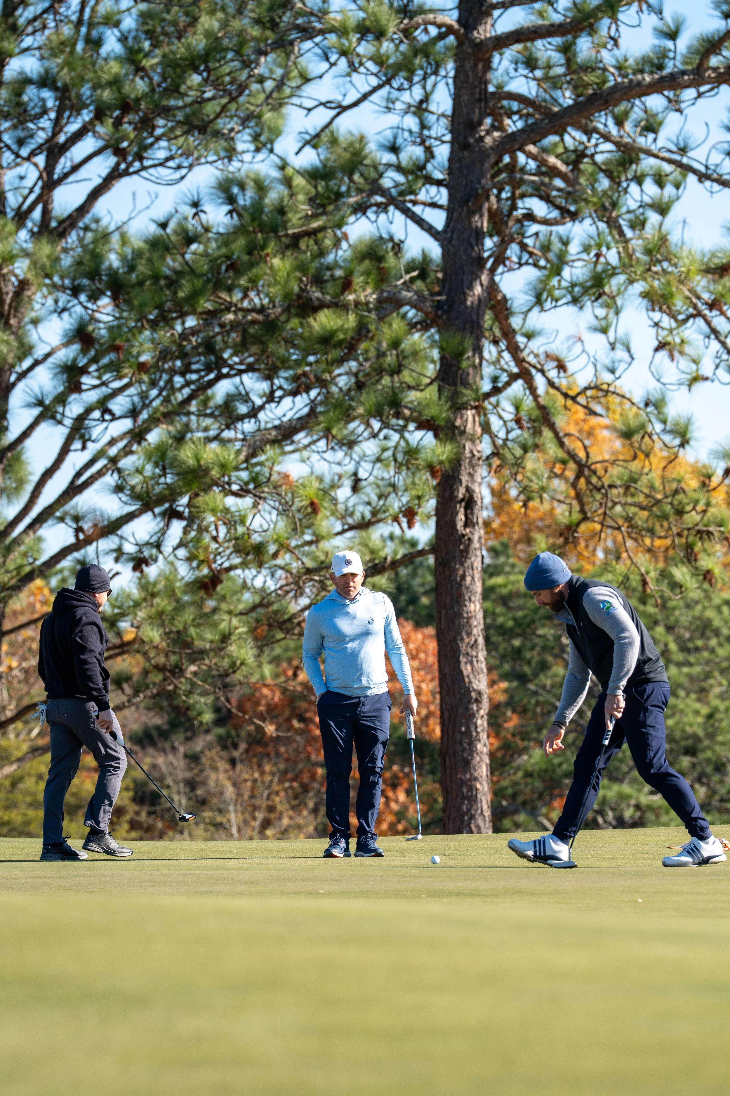Three men playing golf on a green course with tall trees and autumn-colored foliage in the background.
