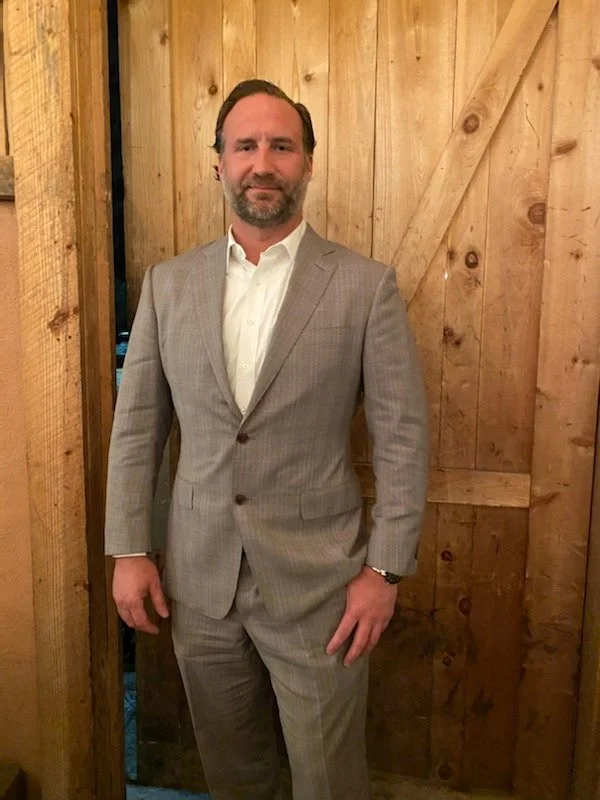 A man with short dark hair and a beard, dressed in a light gray plaid suit and white shirt, standing in front of a wooden wall with diagonal and vertical wood planks.