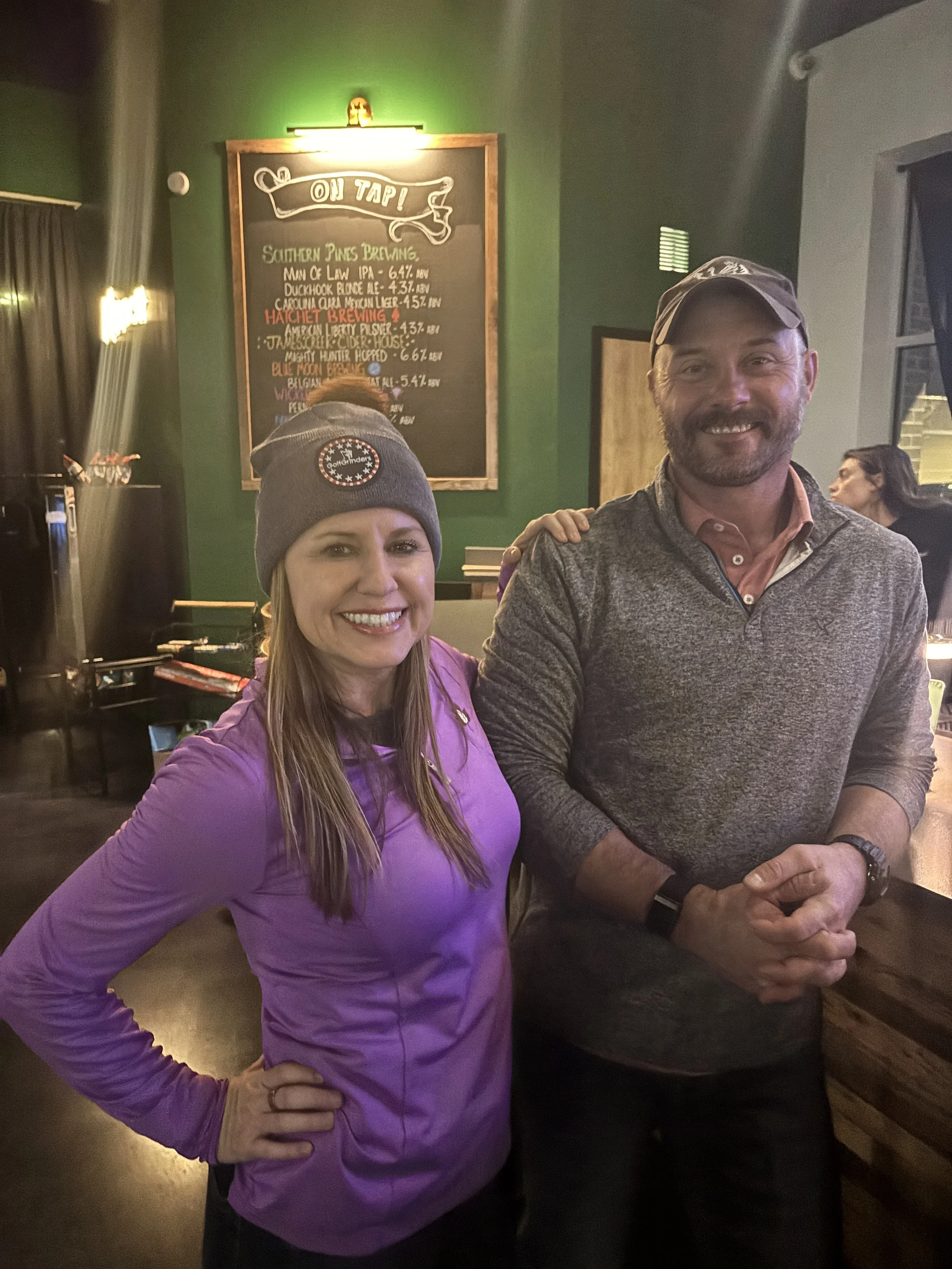 A woman and a man smiling in a bar, with a chalkboard menu behind them listing craft beers.