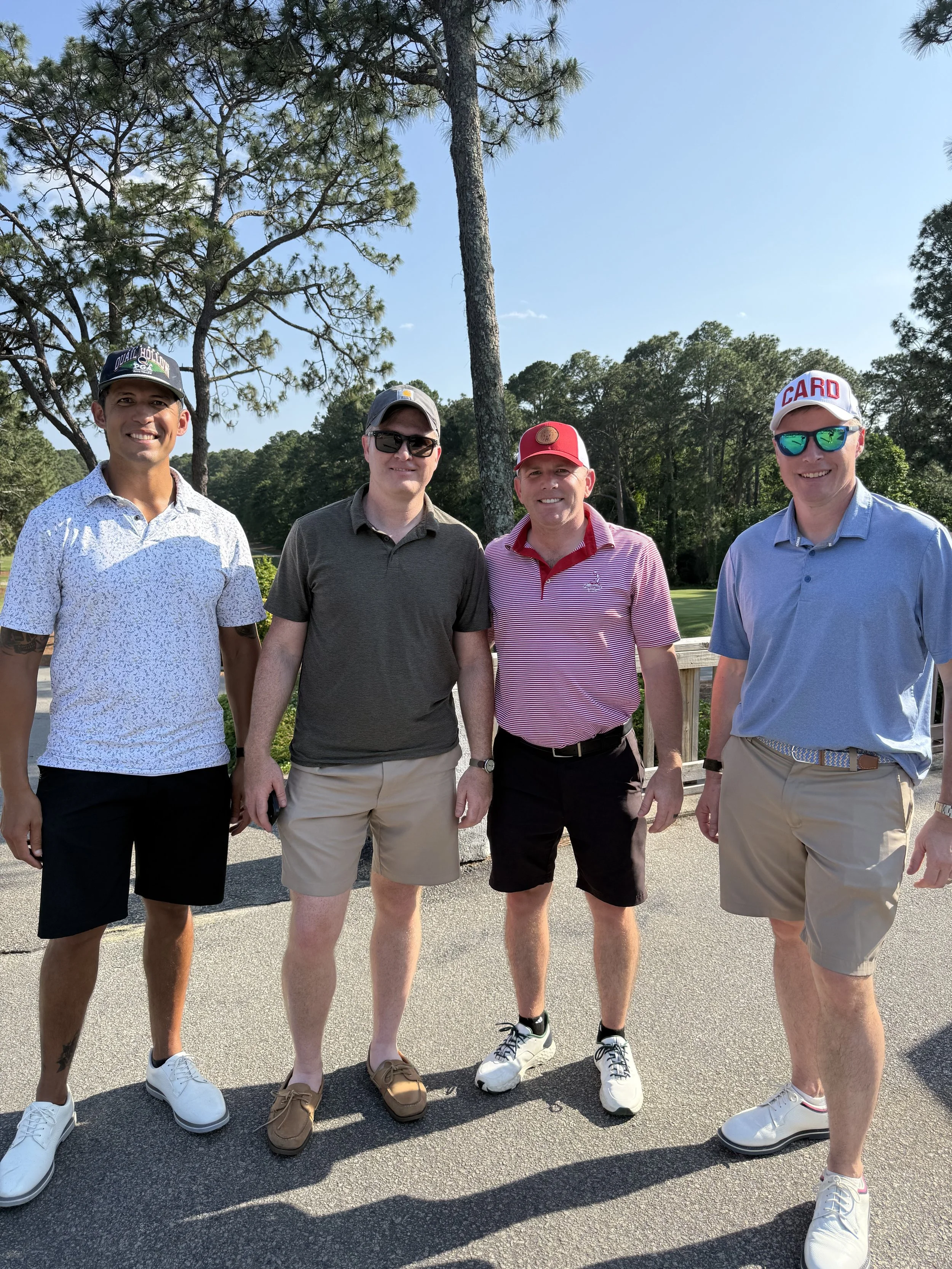 Four men standing together outdoors on a golf course, smiling, wearing golf attire and sunglasses.