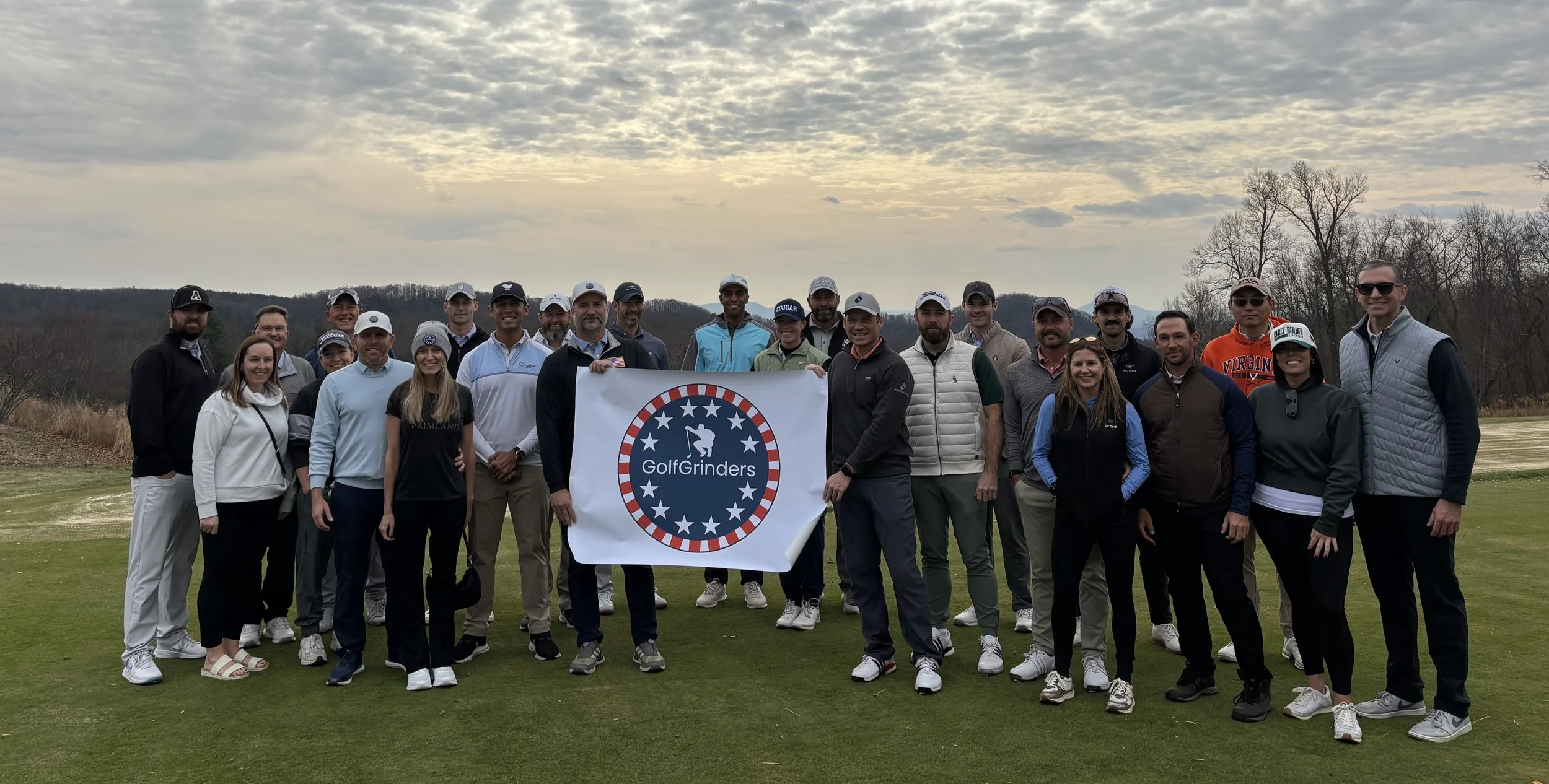 Group of people on a golf course holding a GolfGrinders banner, with trees and cloudy sky in background.