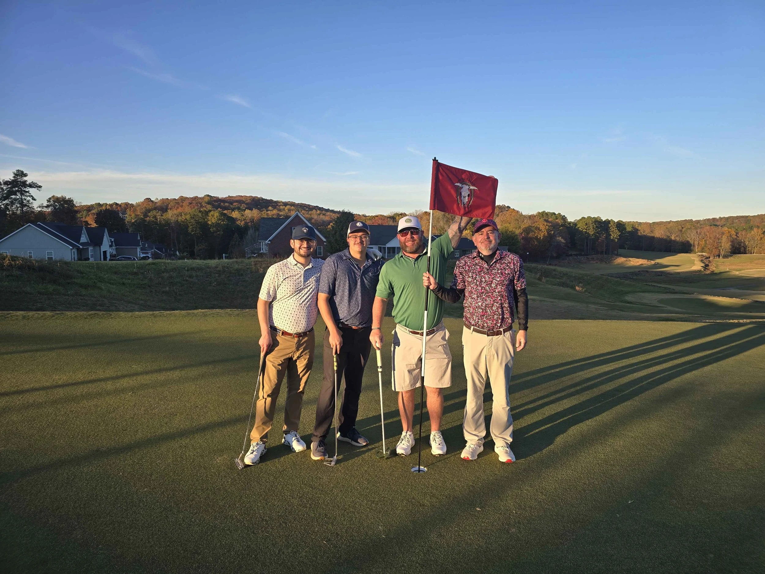 Four men standing on a golf green during sunset, one holding a flag, with houses and hills in the background.
