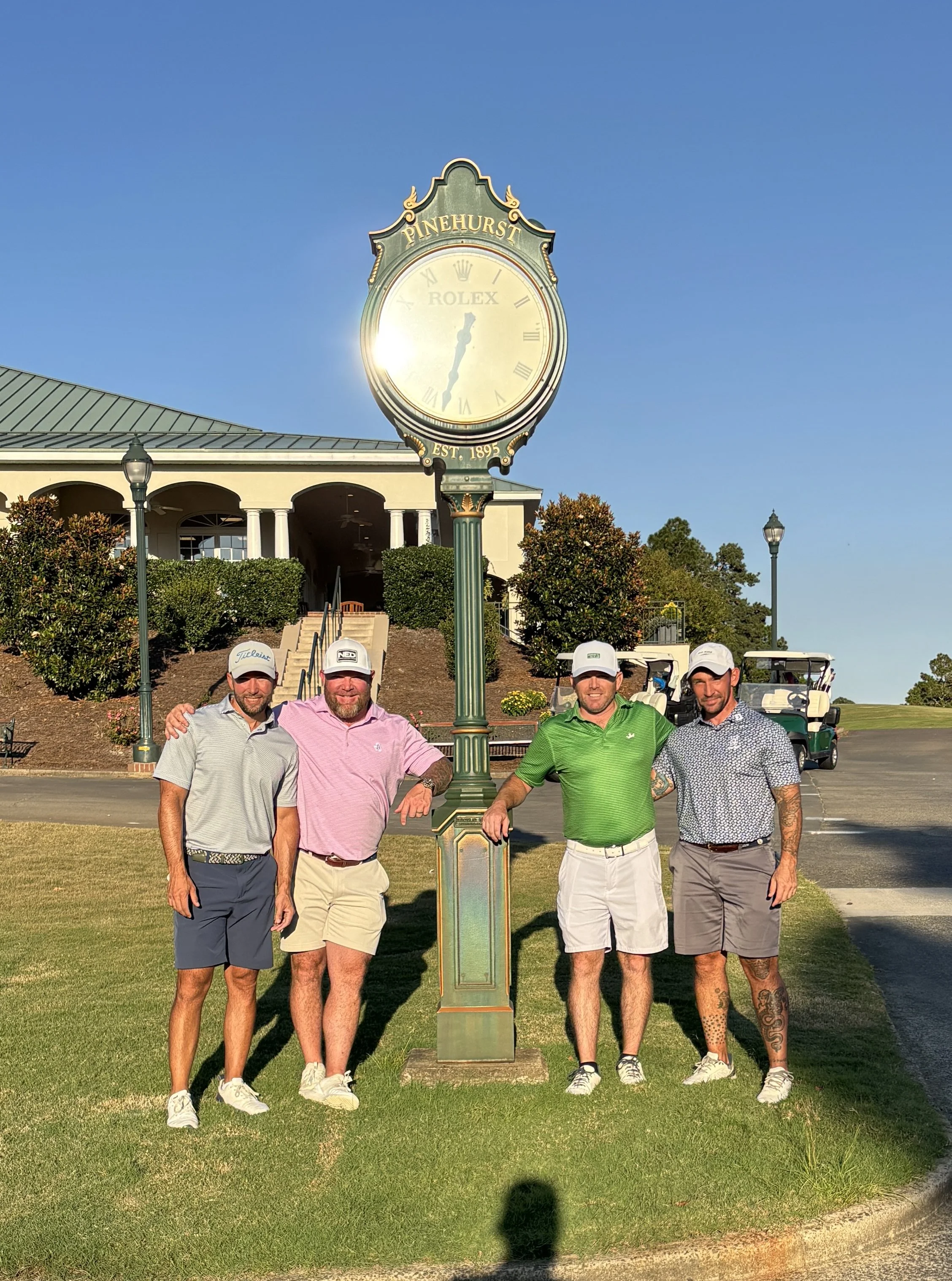 Four men wearing golf attire and holding golf clubs, standing on a golf course near a decorative clock with the words "Pinehurst" and "Rolex" on it, under a clear blue sky.